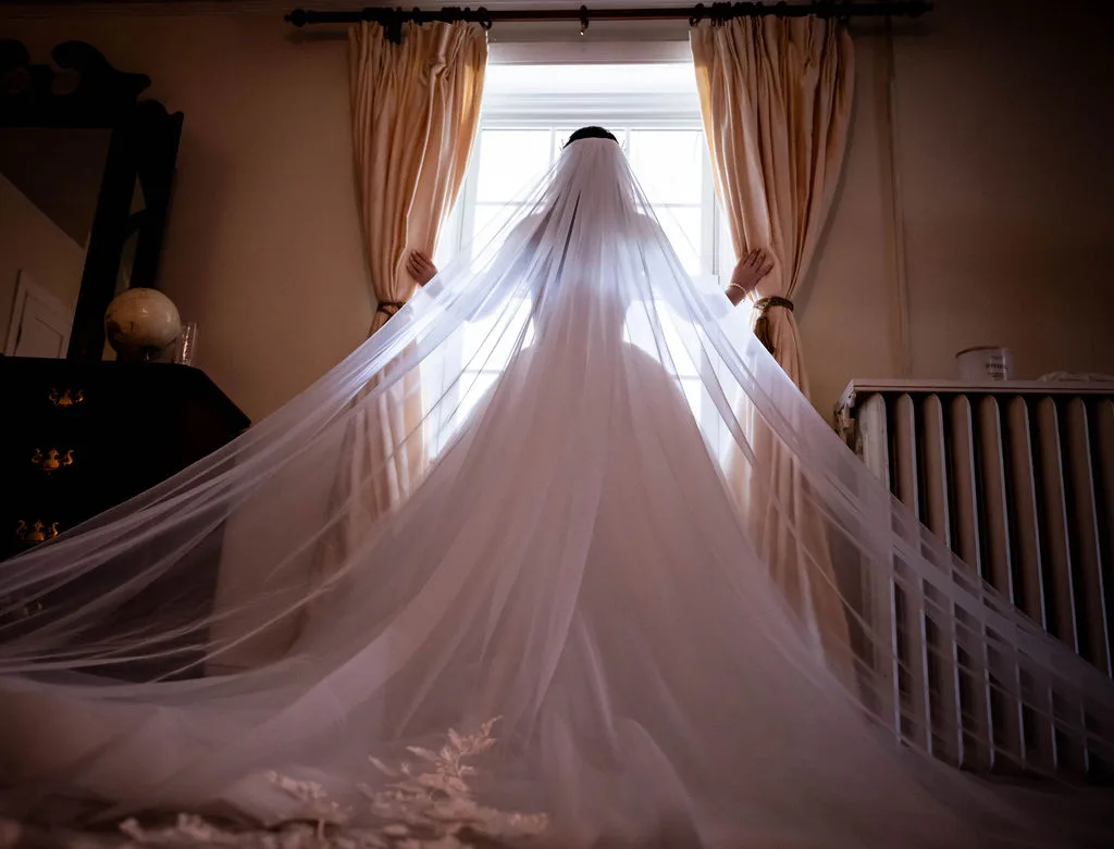 Bride in full cathedral veil and gown stands backlit by window light in a warmly lit dressing room