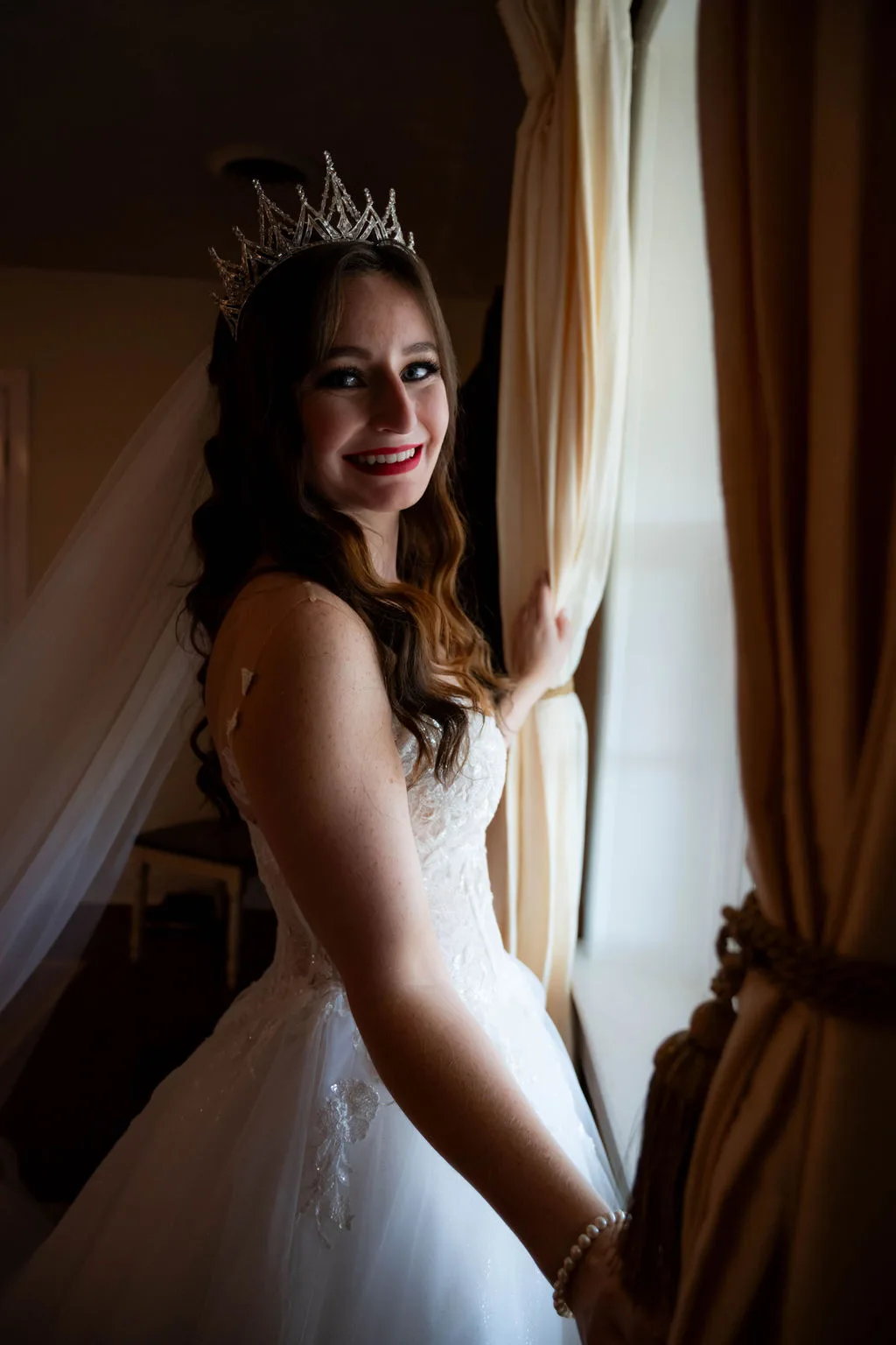 Smiling bride in white gown and crystal tiara poses by draped window curtains before her wedding