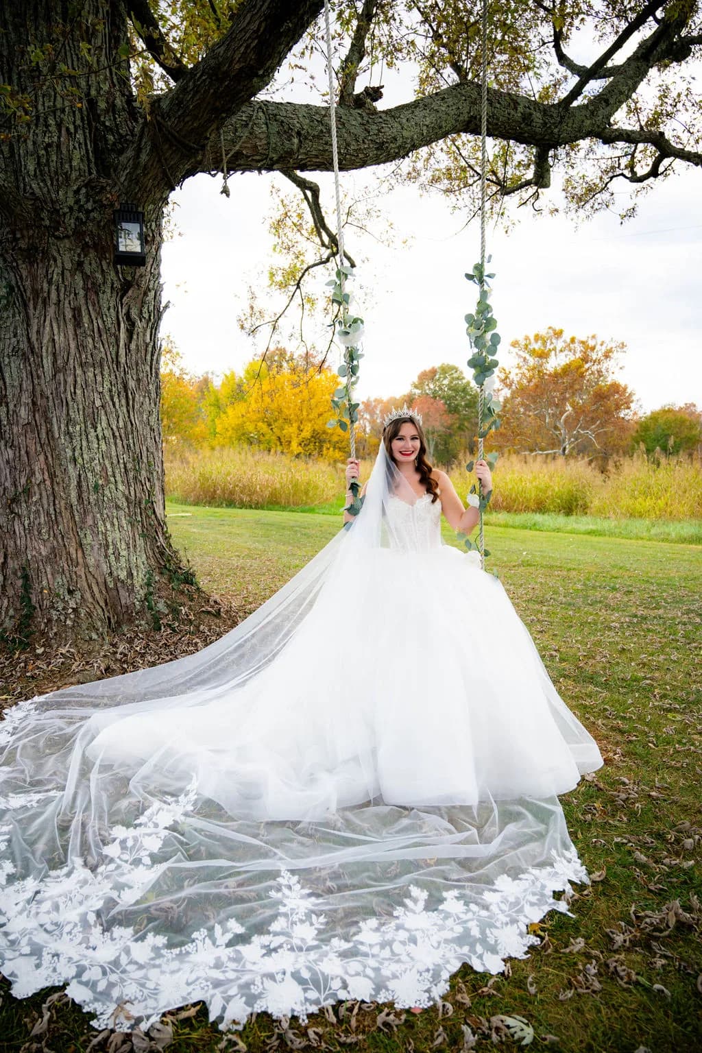 Bride in ballgown sits on eucalyptus-wrapped swing beneath grand oak tree at Rixey Manor in fall foliage