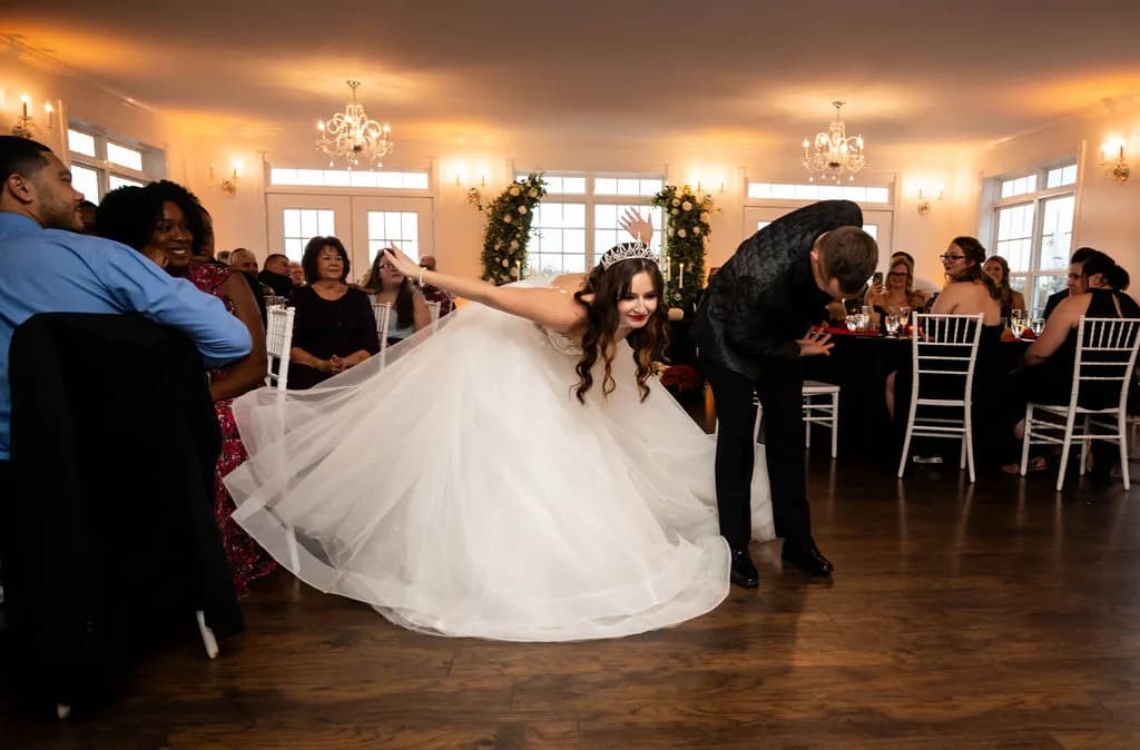Bride in white gown twirls during first dance at elegant indoor reception with guests seated around wooden floor.