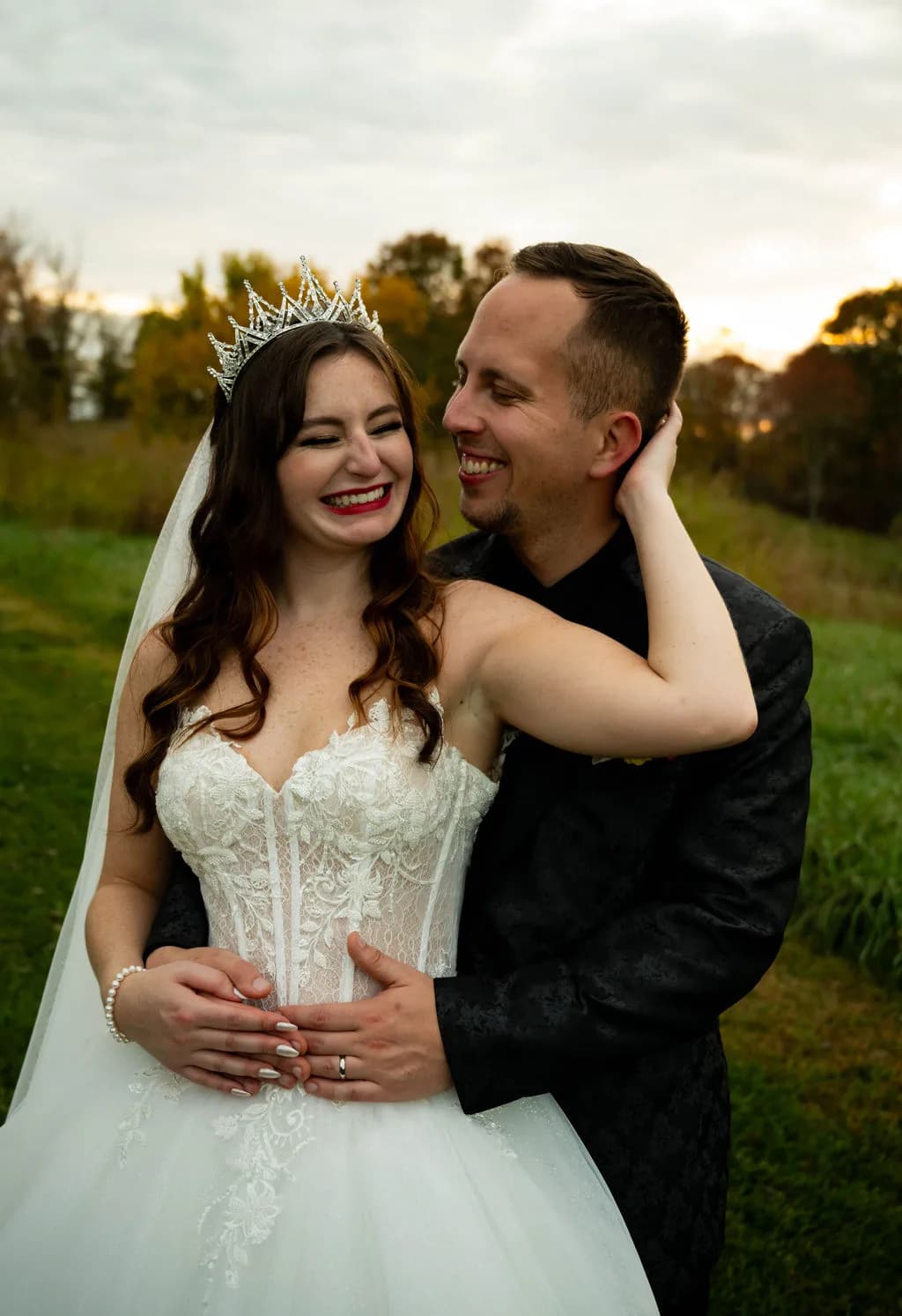 Laughing bride in crown and groom embrace during golden hour portraits on lush green grounds