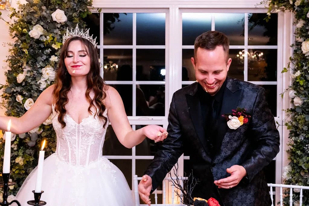 Bride in tiara and groom in black tuxedo laugh together during cake cutting at elegant candlelit reception