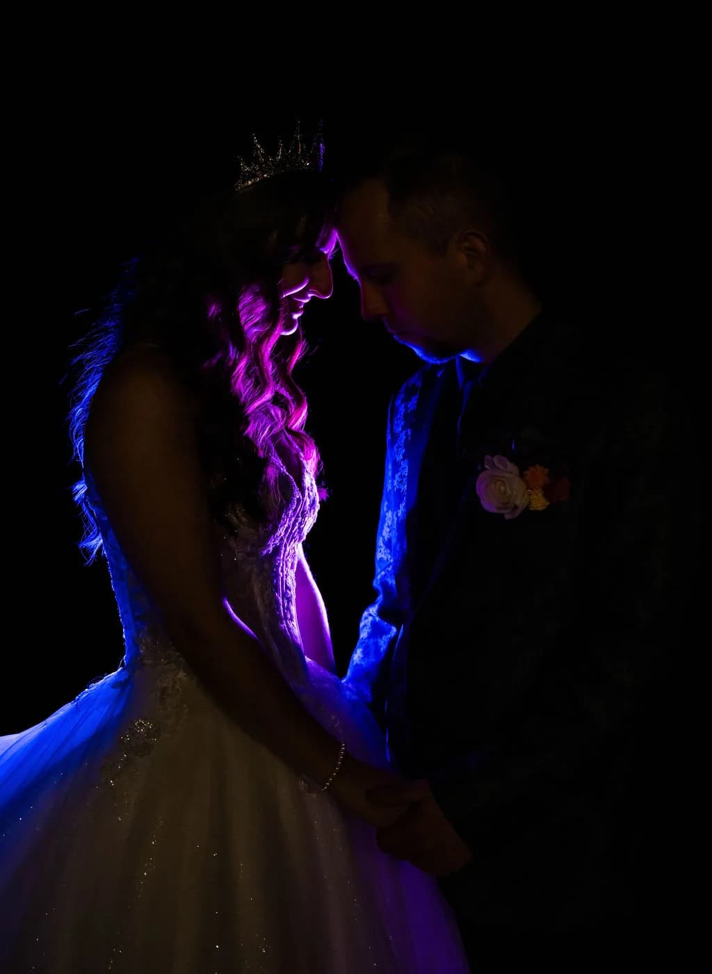 Bride in crown and groom sharing an intimate moment, illuminated by dramatic blue and pink rim lighting against dark background.