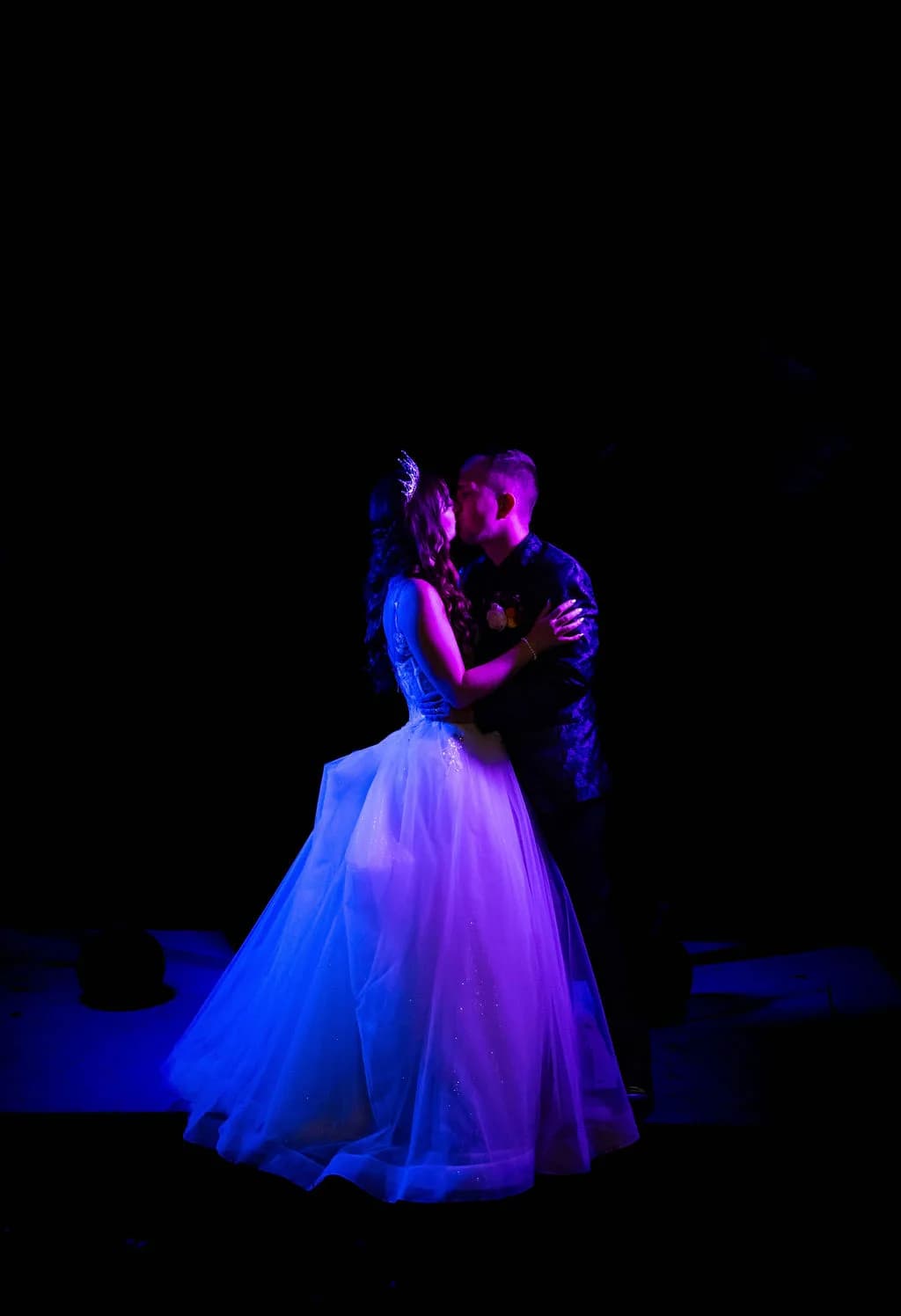 Bride in white ballgown and groom share a kiss during first dance under dramatic blue and purple stage lighting