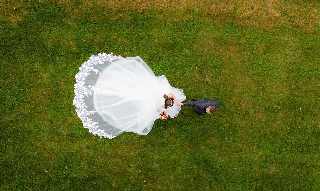 Aerial drone view of bride in sweeping white gown and groom holding bouquet on lush green lawn
