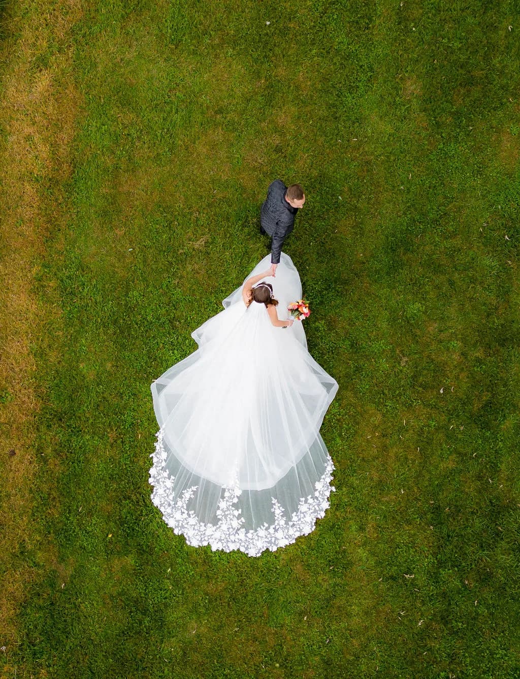 Aerial drone view of bride and groom holding hands on green lawn, her lace-trimmed cathedral veil fanned beautifully below