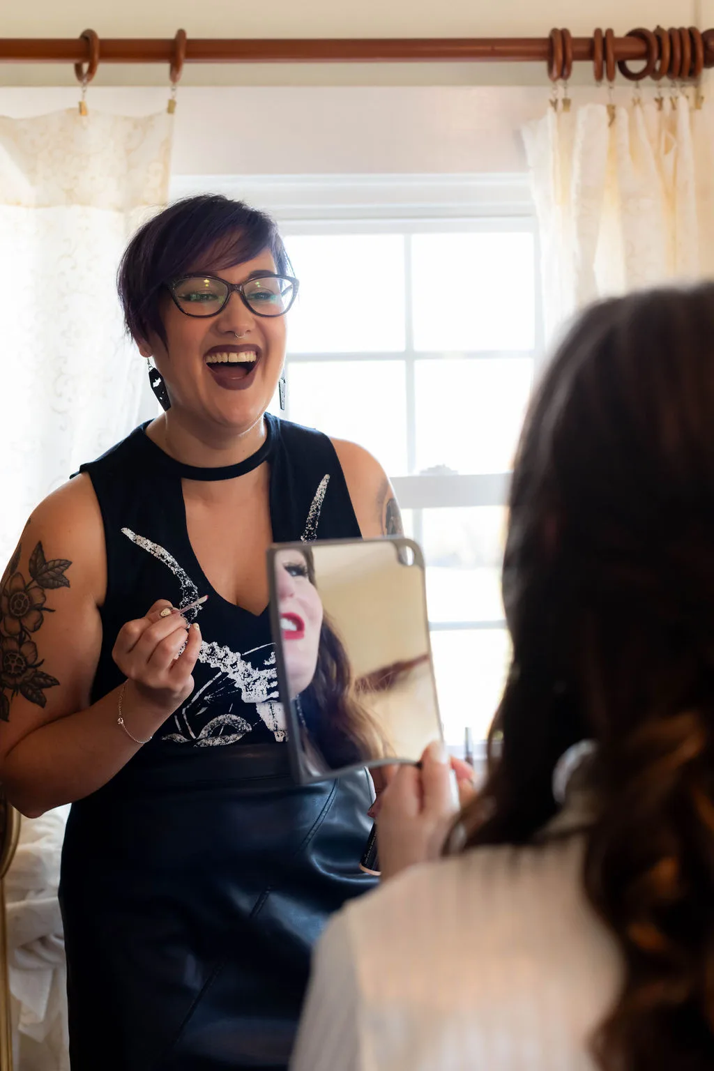 Laughing makeup artist applies mascara to bride holding mirror during wedding morning preparations