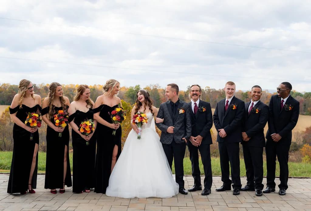 Wedding party in black attire with bride and groom laughing on Rixey Manor terrace with fall foliage backdrop