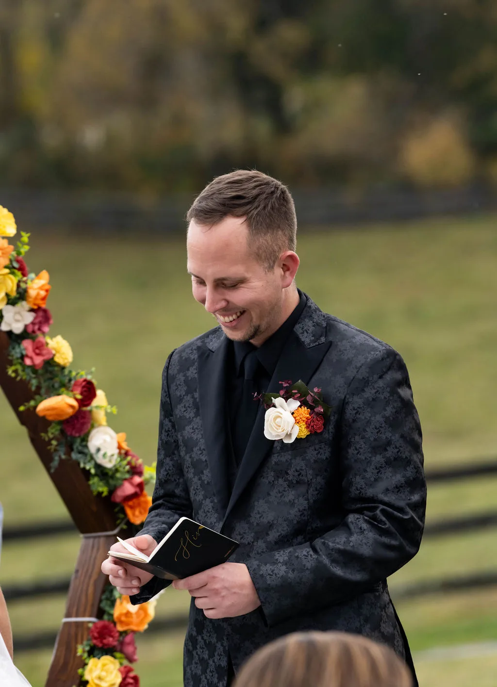 Groom smiling warmly while reading vows at outdoor fall ceremony beside floral arch at Rixey Manor