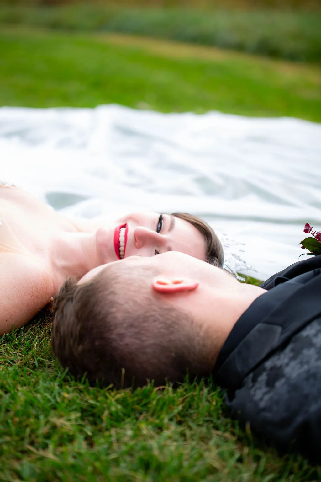 Bride and groom lying on grass beneath wedding veil, smiling up at camera on Rixey Manor grounds
