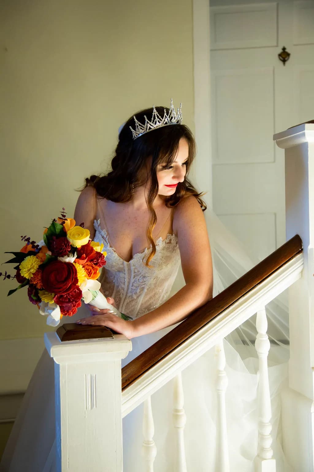 Bride in lace gown and crystal tiara holding vibrant autumn bouquet on a white staircase railing