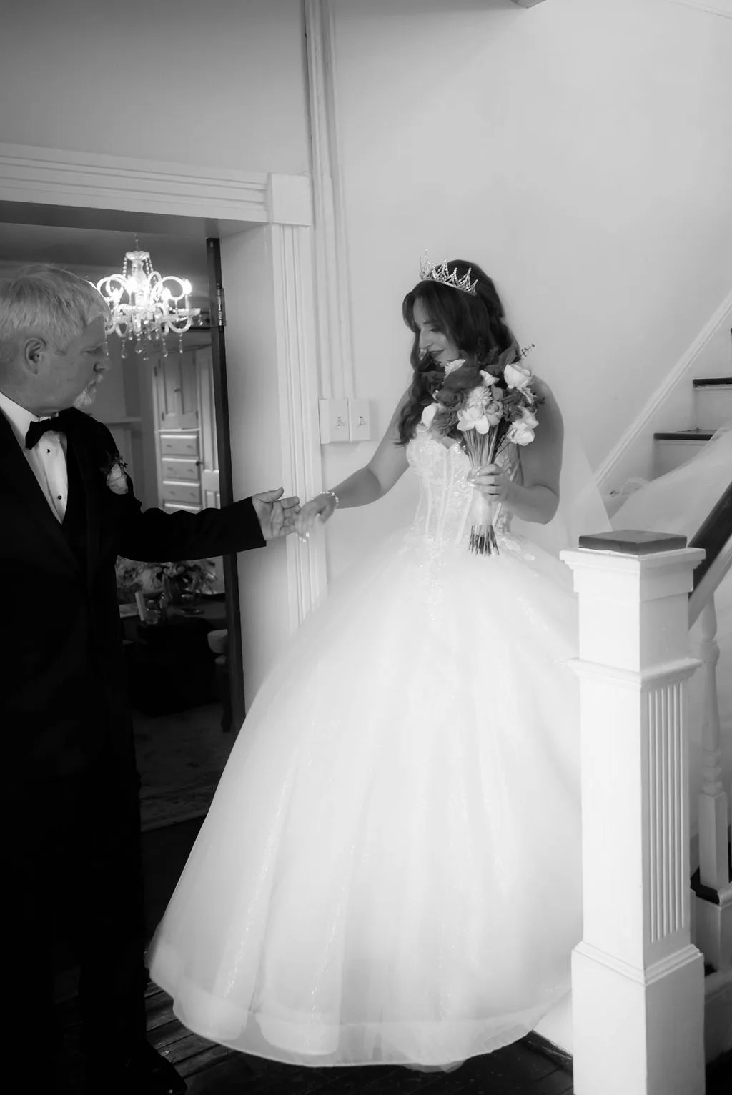 Bride in tiara and ball gown descends staircase holding bouquet, escorted by man in tuxedo