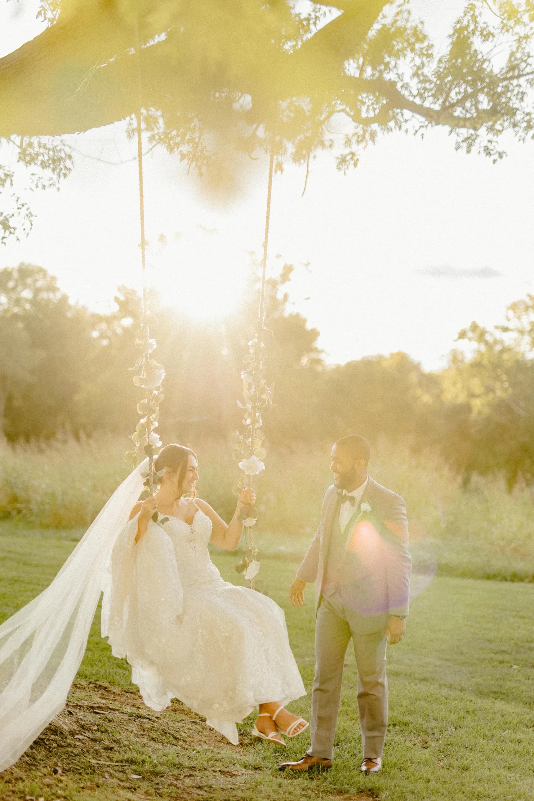 Bride on floral swing under golden sunset light as groom looks on adoringly at Rixey Manor grounds