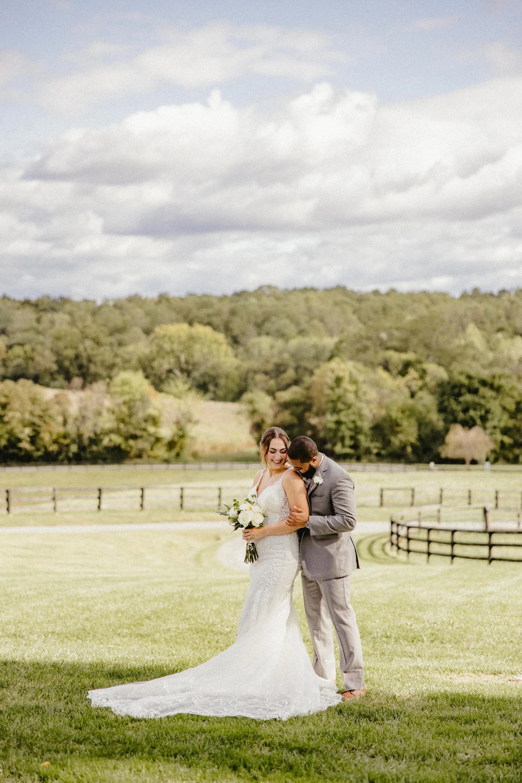 Groom embraces bride from behind on Rixey Manor's sunlit lawn with rolling hills and pastoral fencing behind them.