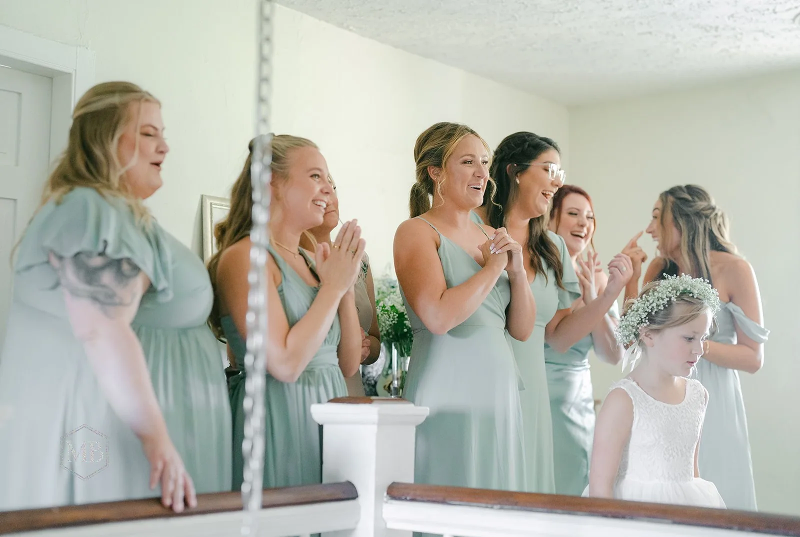 Bridesmaids in sage dresses react with joy and clapping as flower girl looks on during wedding preparations