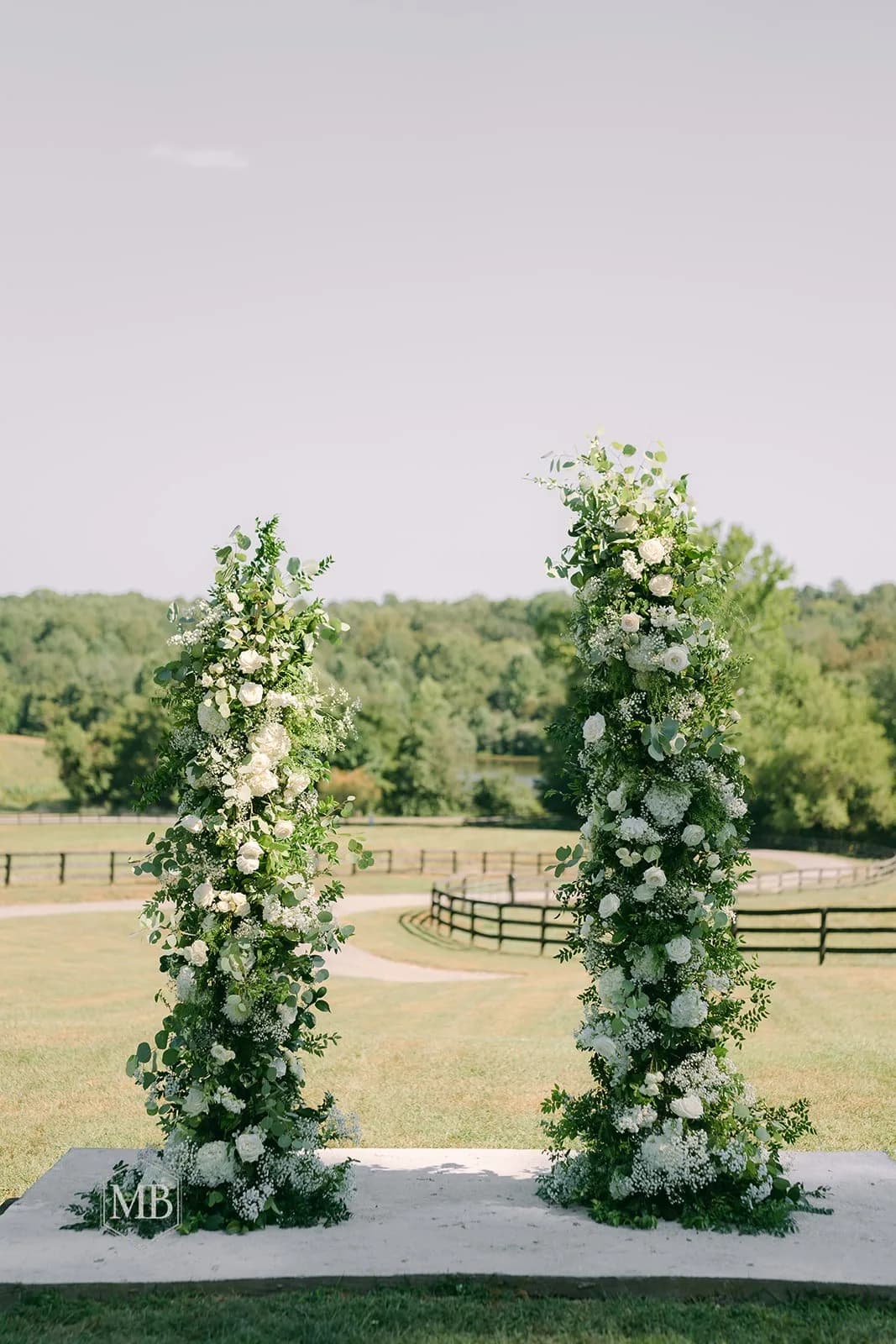 Two lush floral columns wrapped in white blooms and greenery at an outdoor ceremony site overlooking Rixey Manor's pastoral grounds