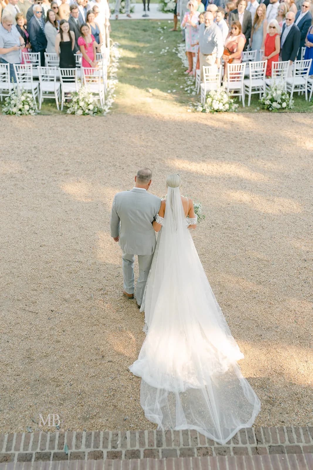 Aerial view of bride and escort walking down outdoor aisle at Rixey Manor, guests lining white chairs