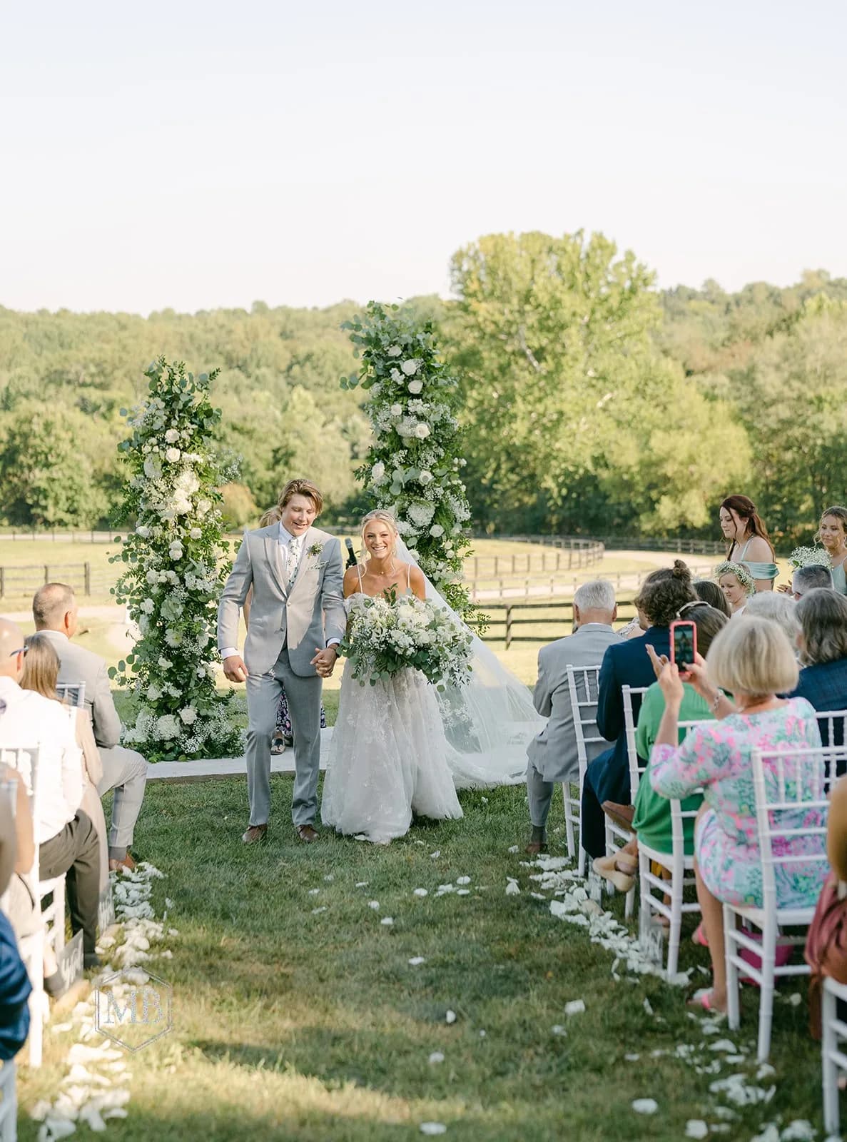 Bride and groom walking down aisle at outdoor western wedding ceremony with green arch and guests seated on both sides