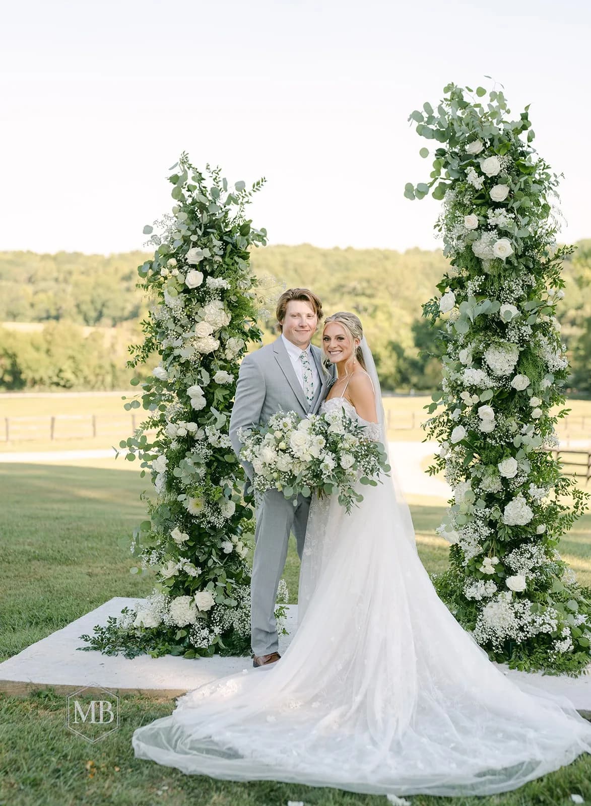 Bride and groom smile between two towering floral columns with white roses and greenery on sunny Virginia estate grounds.