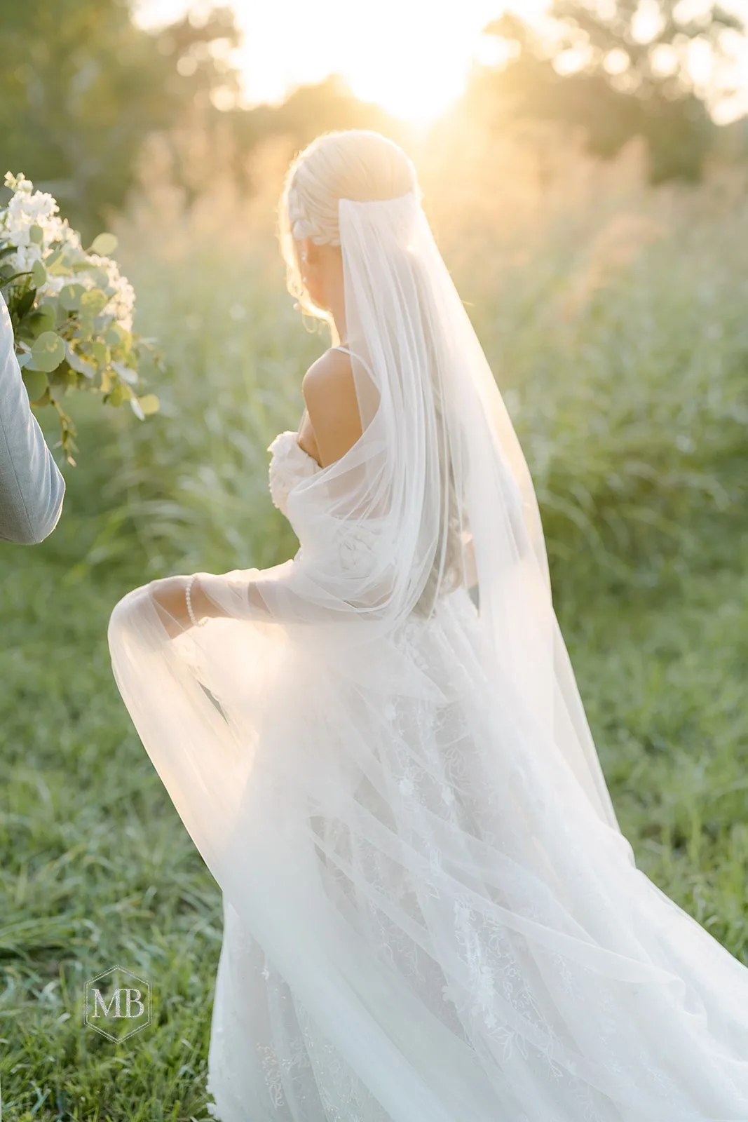 Bride in white gown and veil holding bouquet in sunlit garden field at golden hour