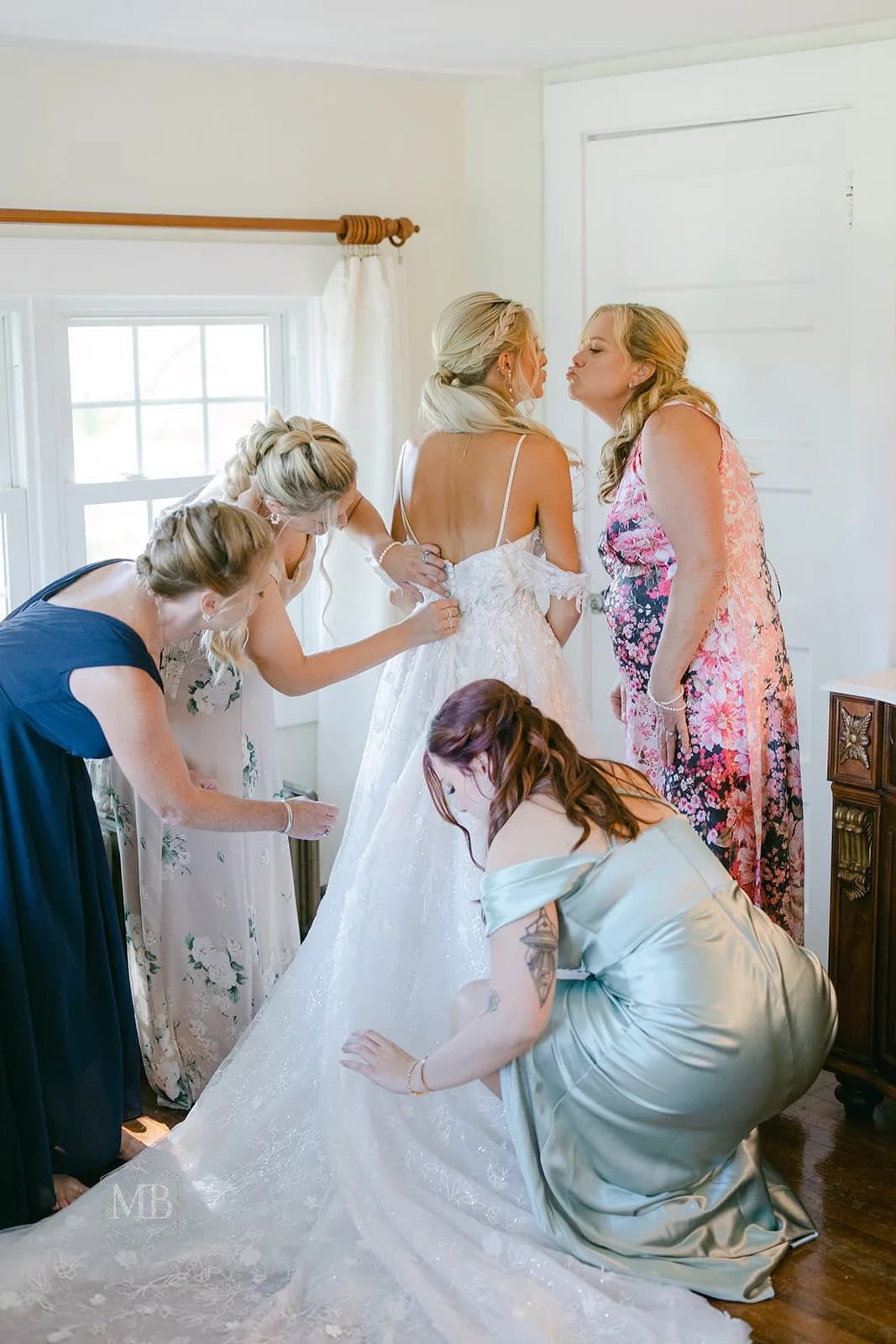 Bridesmaids helping bride into her gown, one giving a kiss on the cheek, in a sunlit getting-ready room