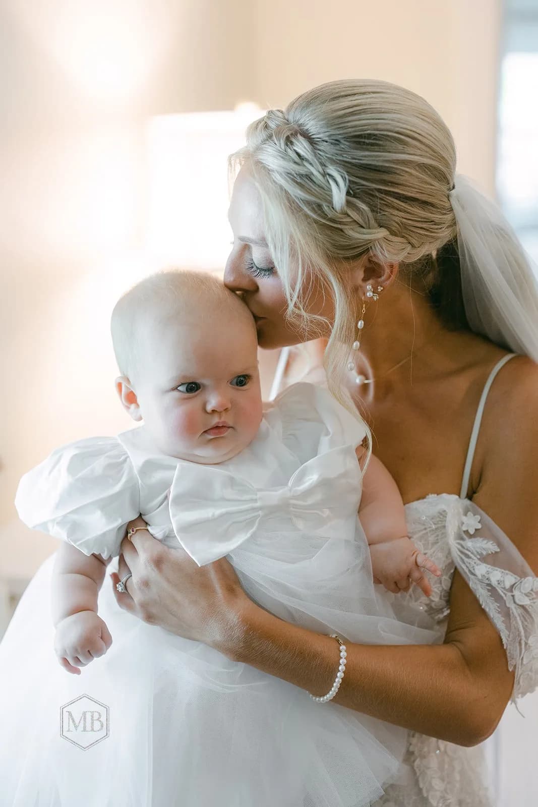 Bride in white lace gown kisses infant in satin bow dress during wedding day getting-ready moment