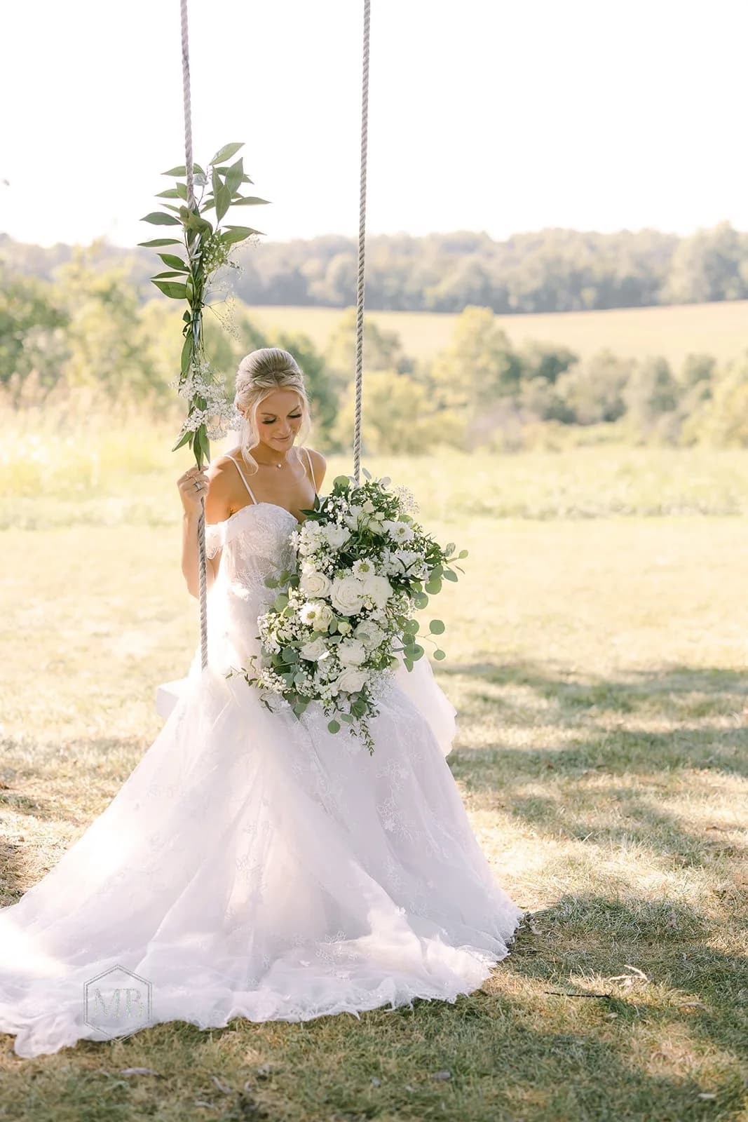 Bride in flowing gown sits on rope swing holding lush white bouquet on sunlit Virginia estate grounds