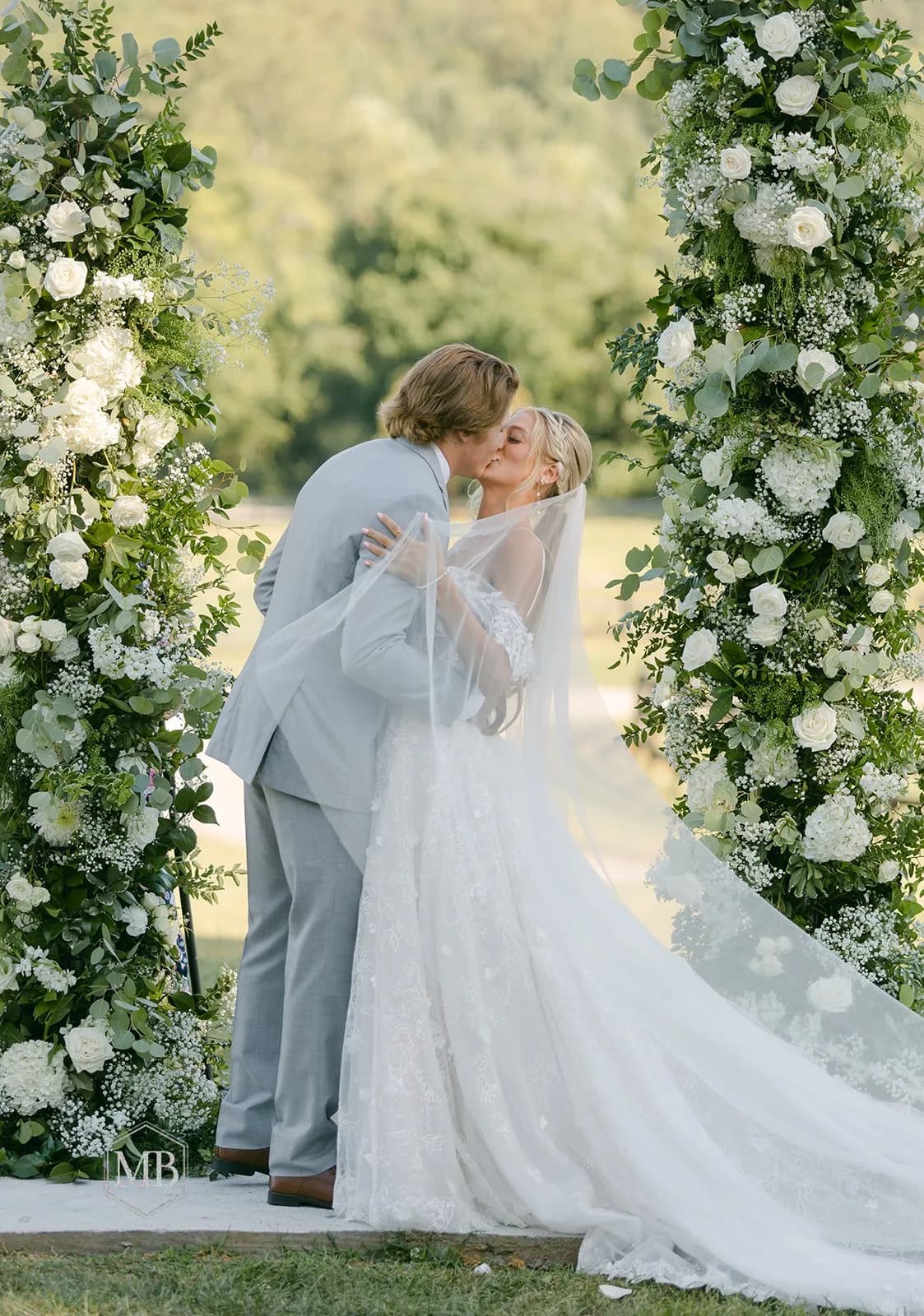Bride and groom share first kiss under lush white floral arch at outdoor Rixey Manor ceremony