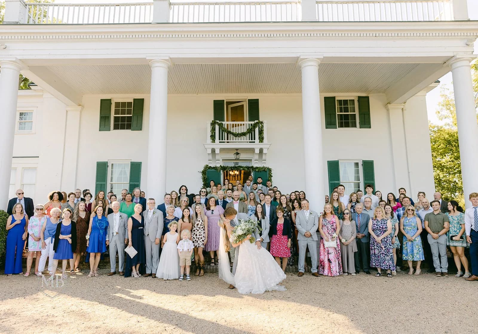 Bride and groom share a kiss before full wedding guest group portrait at Rixey Manor's white columned facade
