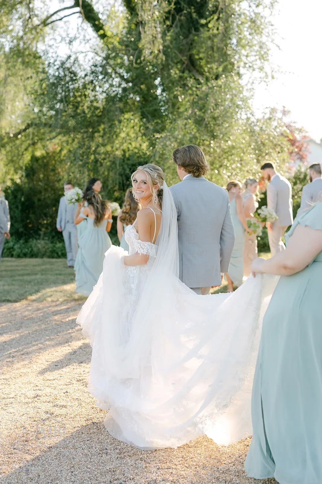 Bride glances back smiling as wedding party walks the sun-dappled grounds at Rixey Manor