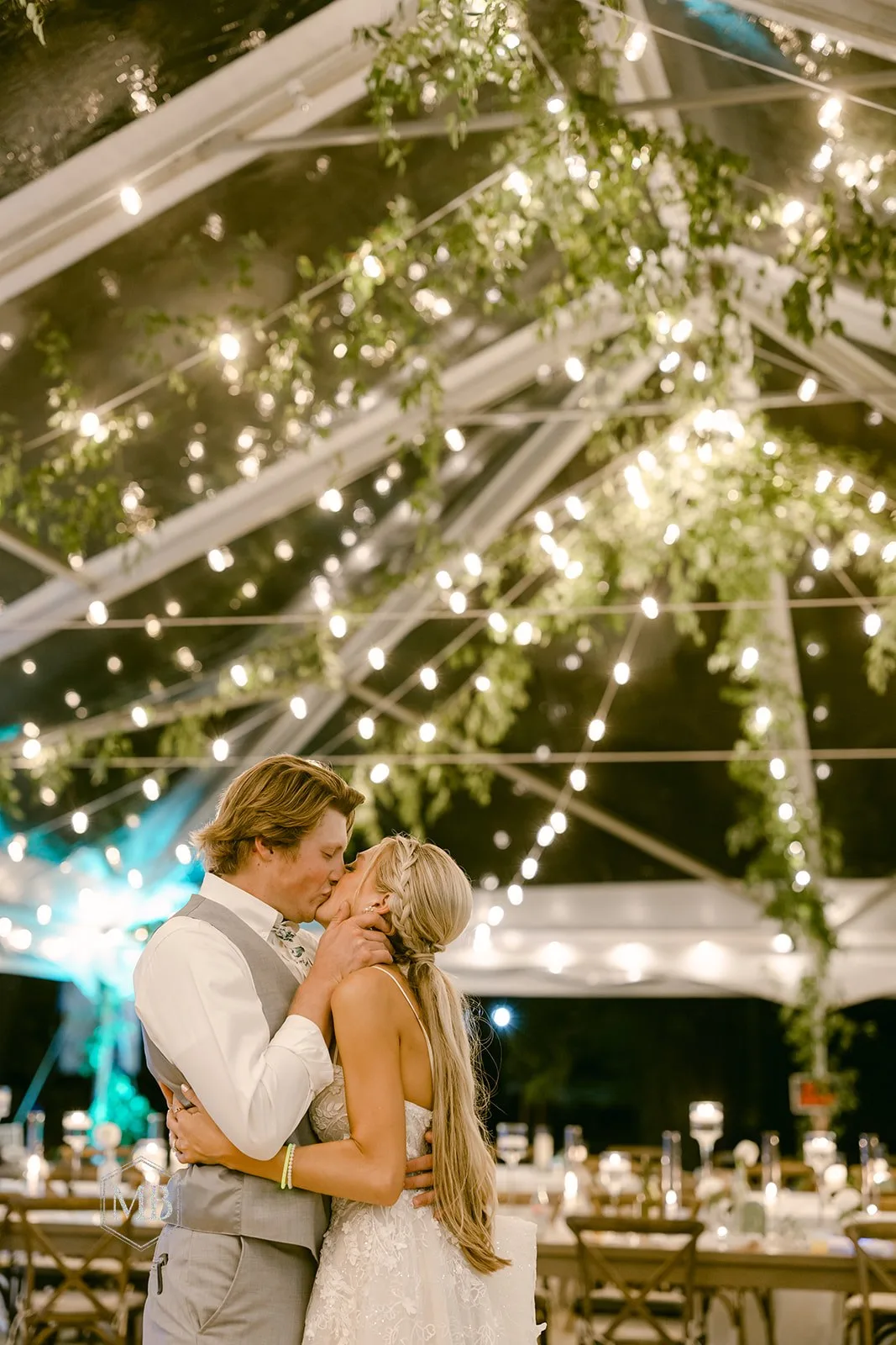 Bride and groom share a kiss under a glowing tent draped with string lights and greenery at a evening reception.