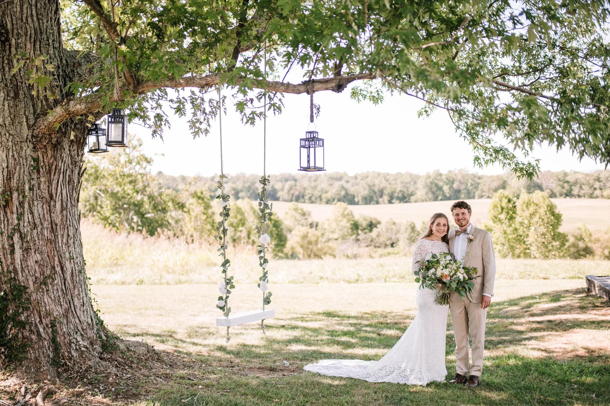 Bride and groom portrait under large oak tree with hanging lantern and scenic field backdrop