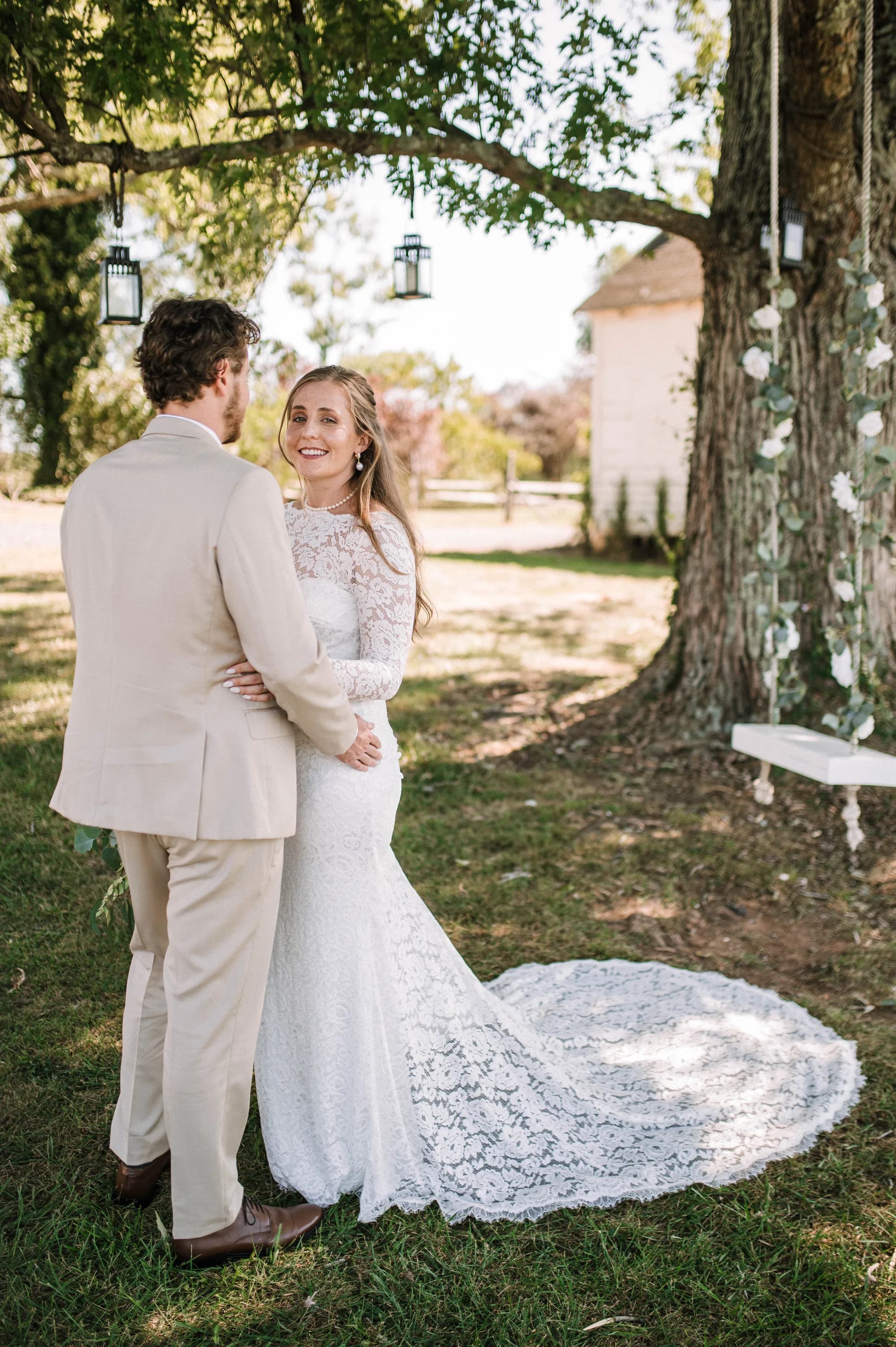 Bride in lace gown smiles over shoulder embracing groom under lantern-hung tree at Rixey Manor grounds