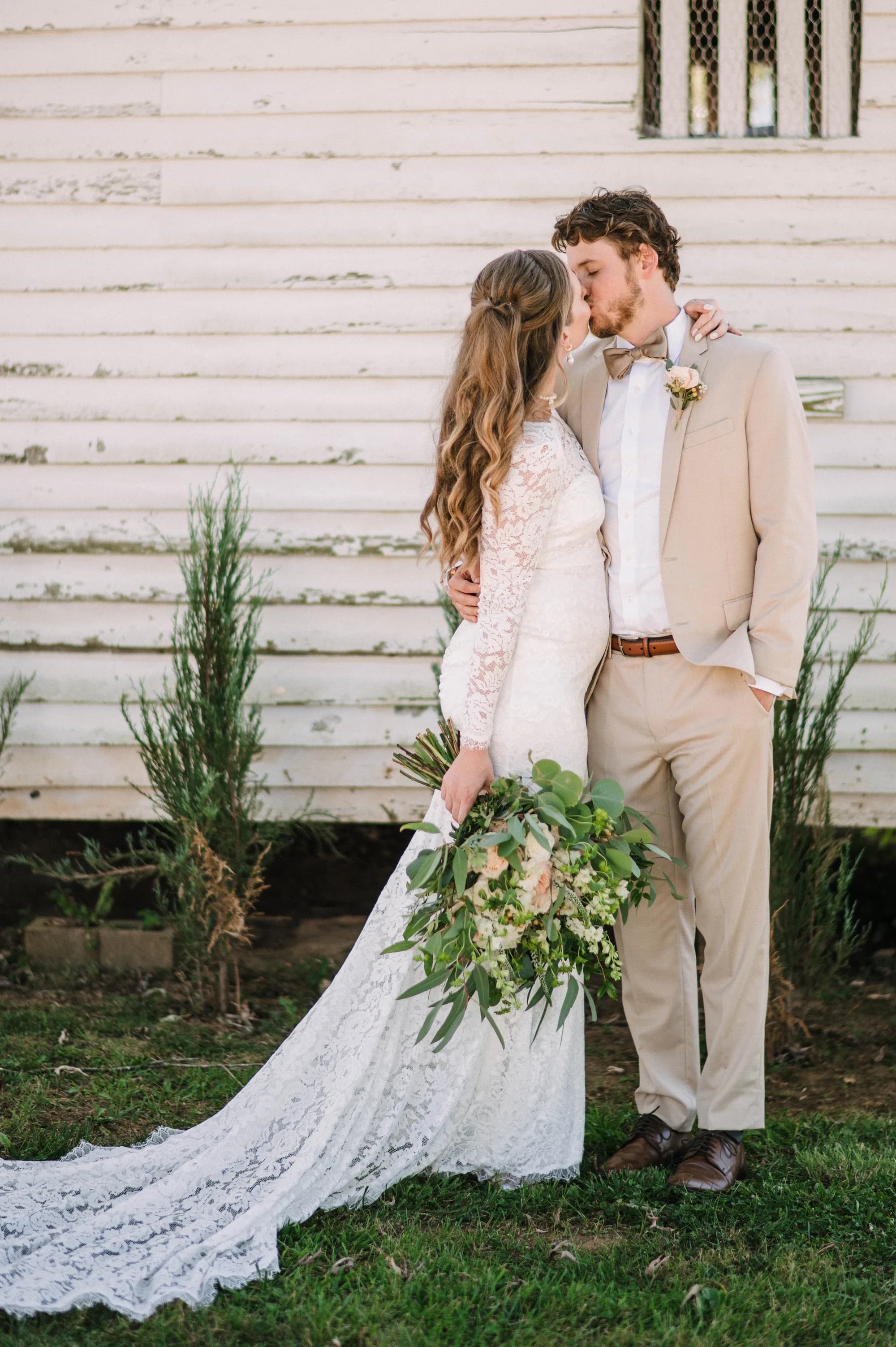 Bride and groom share a kiss outside a white farmhouse, bride holding lush greenery bouquet in lace gown