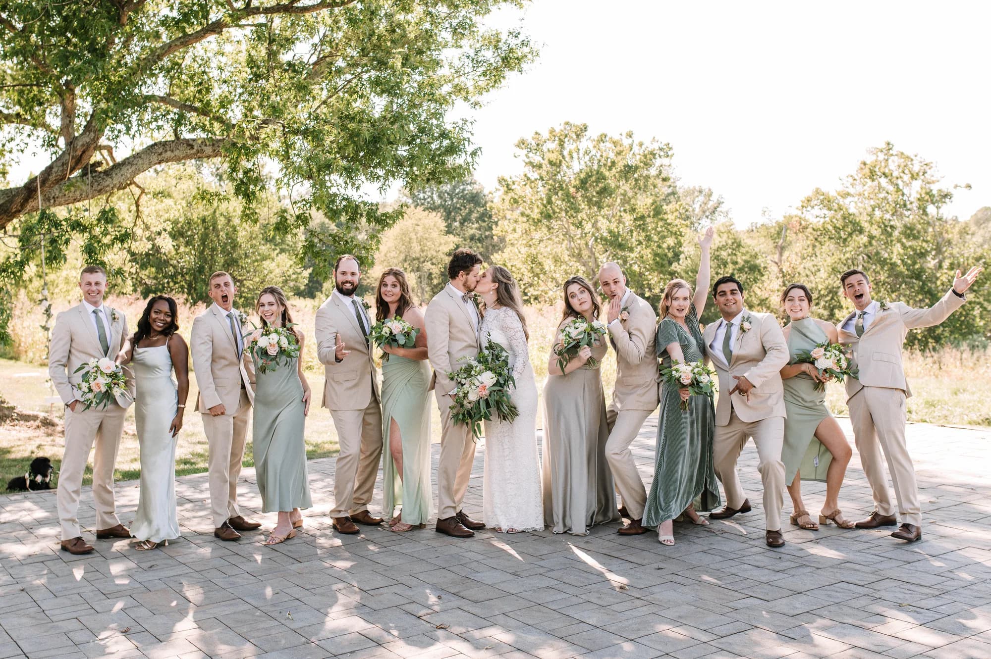Wedding party in sage and tan attire laughs and celebrates as couple kisses outdoors at Rixey Manor