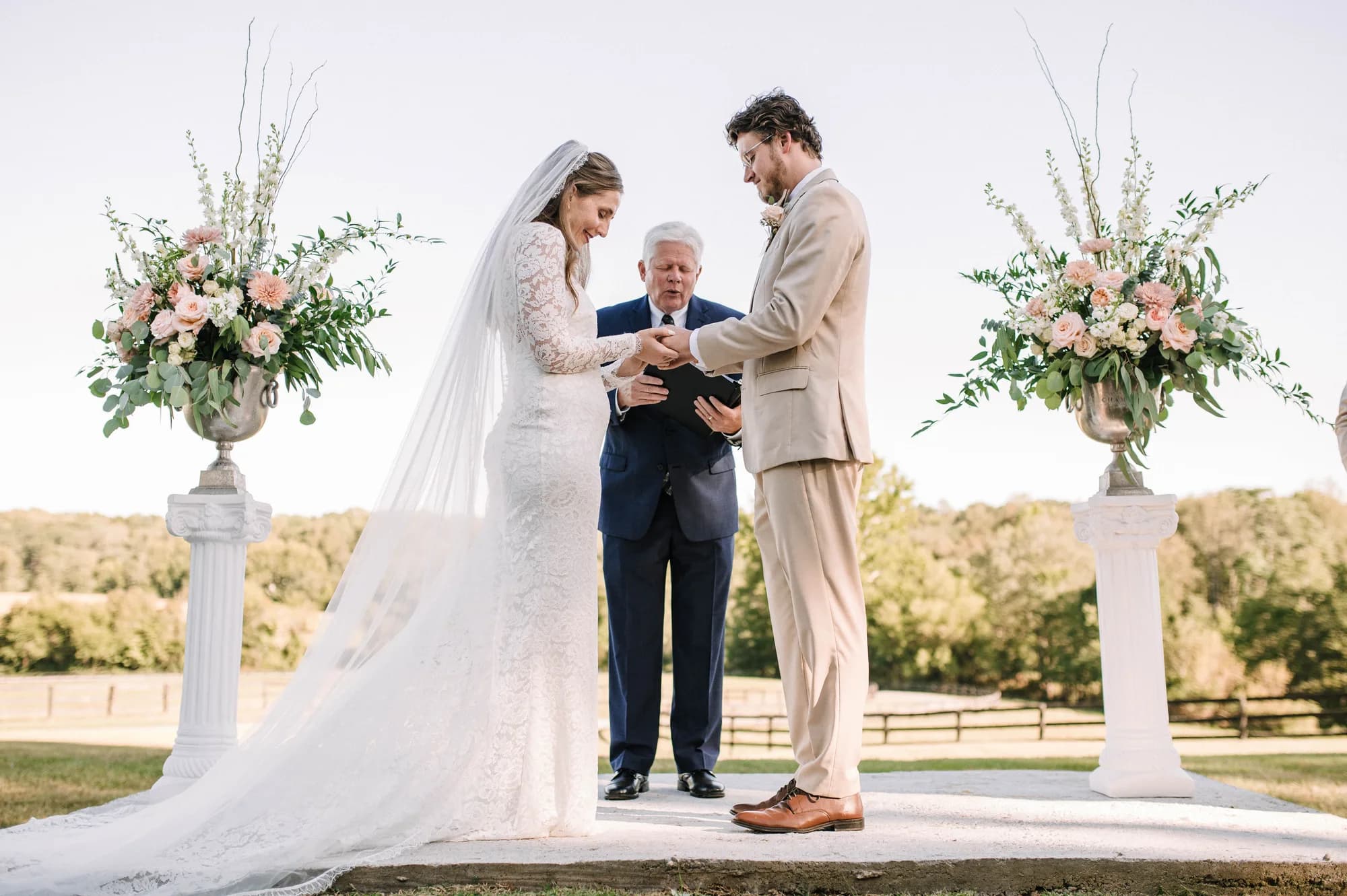 Bride and groom exchange rings at outdoor Rixey Manor ceremony flanked by lush floral urns on white pedestals