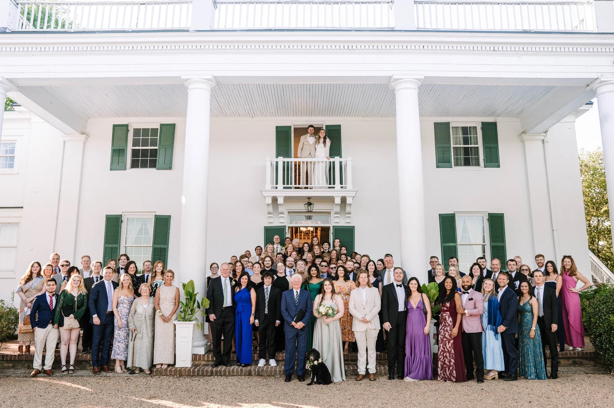 Full wedding group portrait in front of Rixey Manor's white columned facade with couple on balcony above