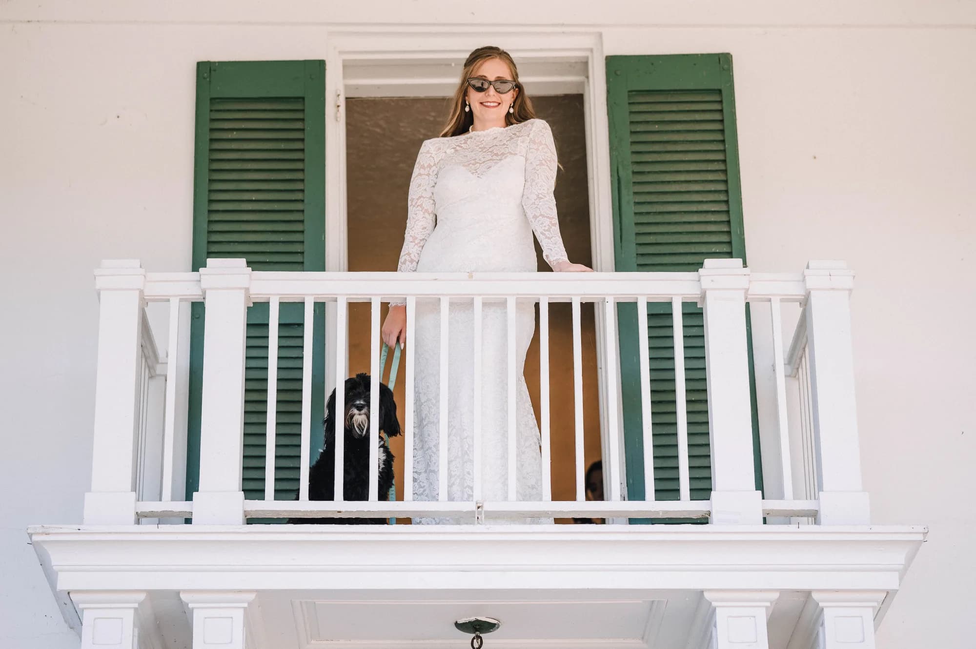 Smiling bride in white lace gown stands on white balcony with green shutters at Rixey Manor with dog