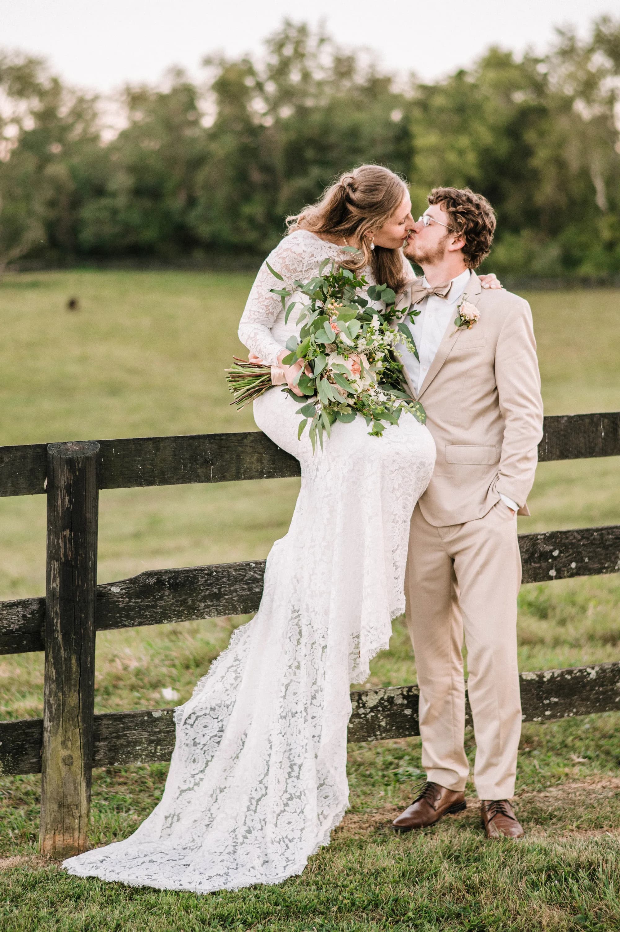 Bride and groom kiss by wooden fence in pastoral setting with green fields and trees behind them