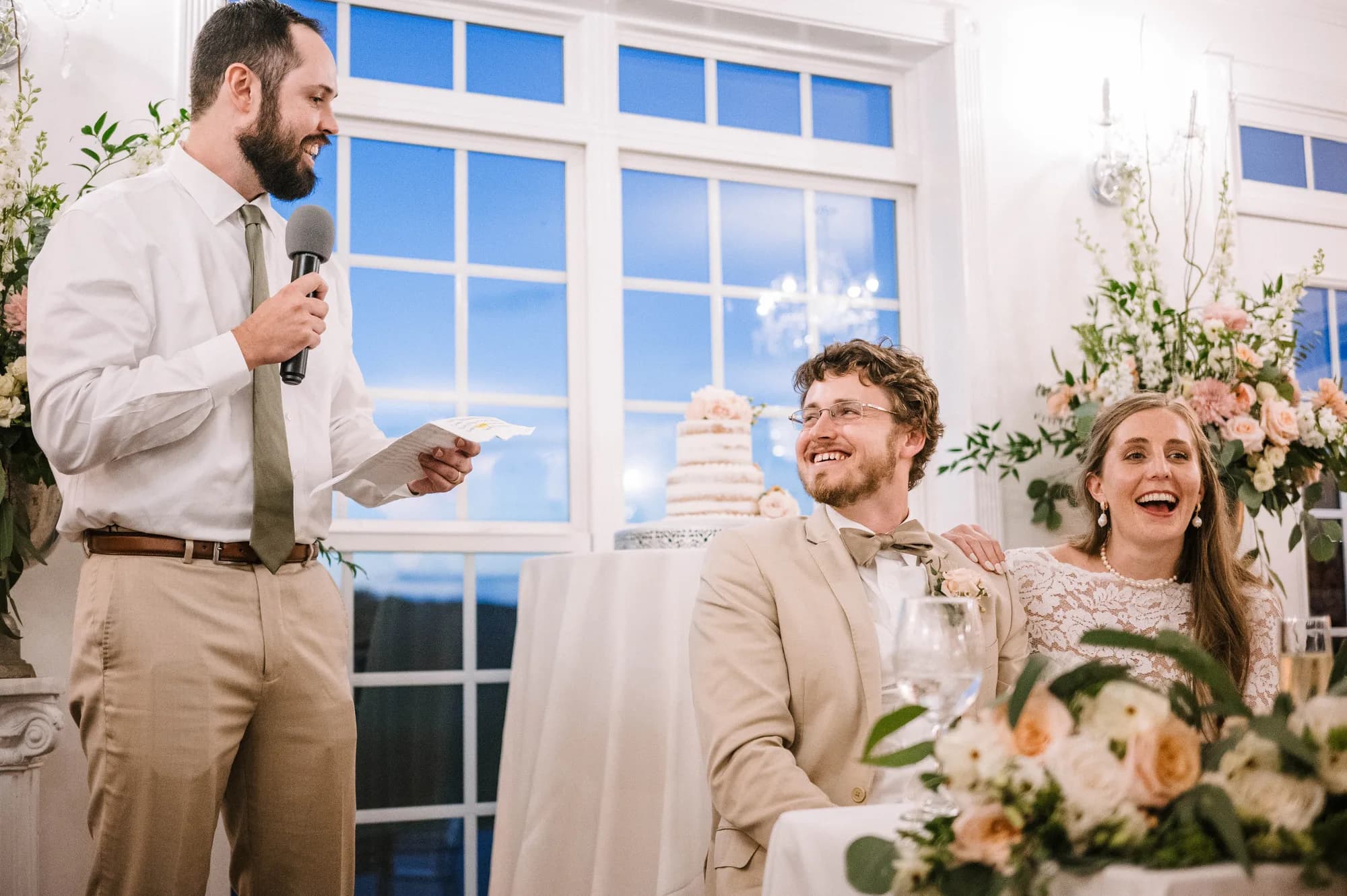 Best man delivers toast as bride and groom laugh joyfully at wedding reception table with floral arrangements