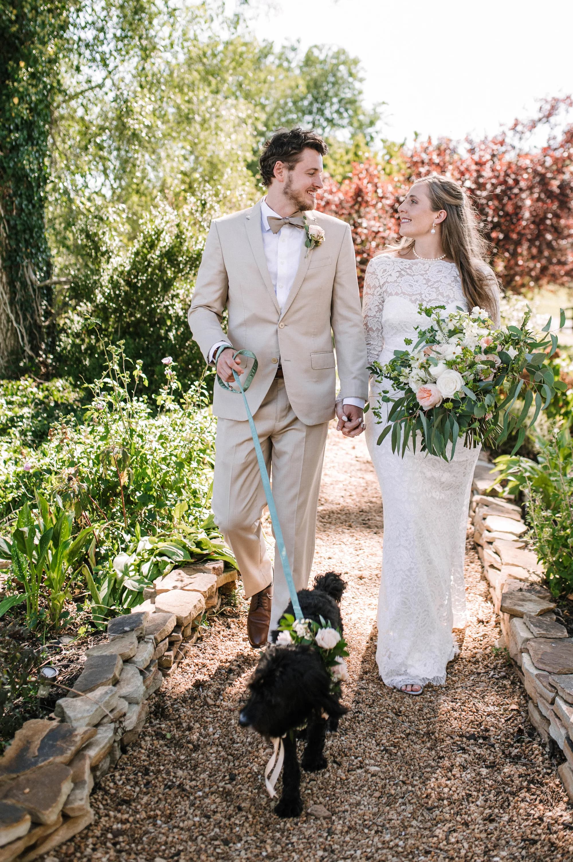 Bride and groom walk hand-in-hand along a sunlit garden path with their black dog at Rixey Manor