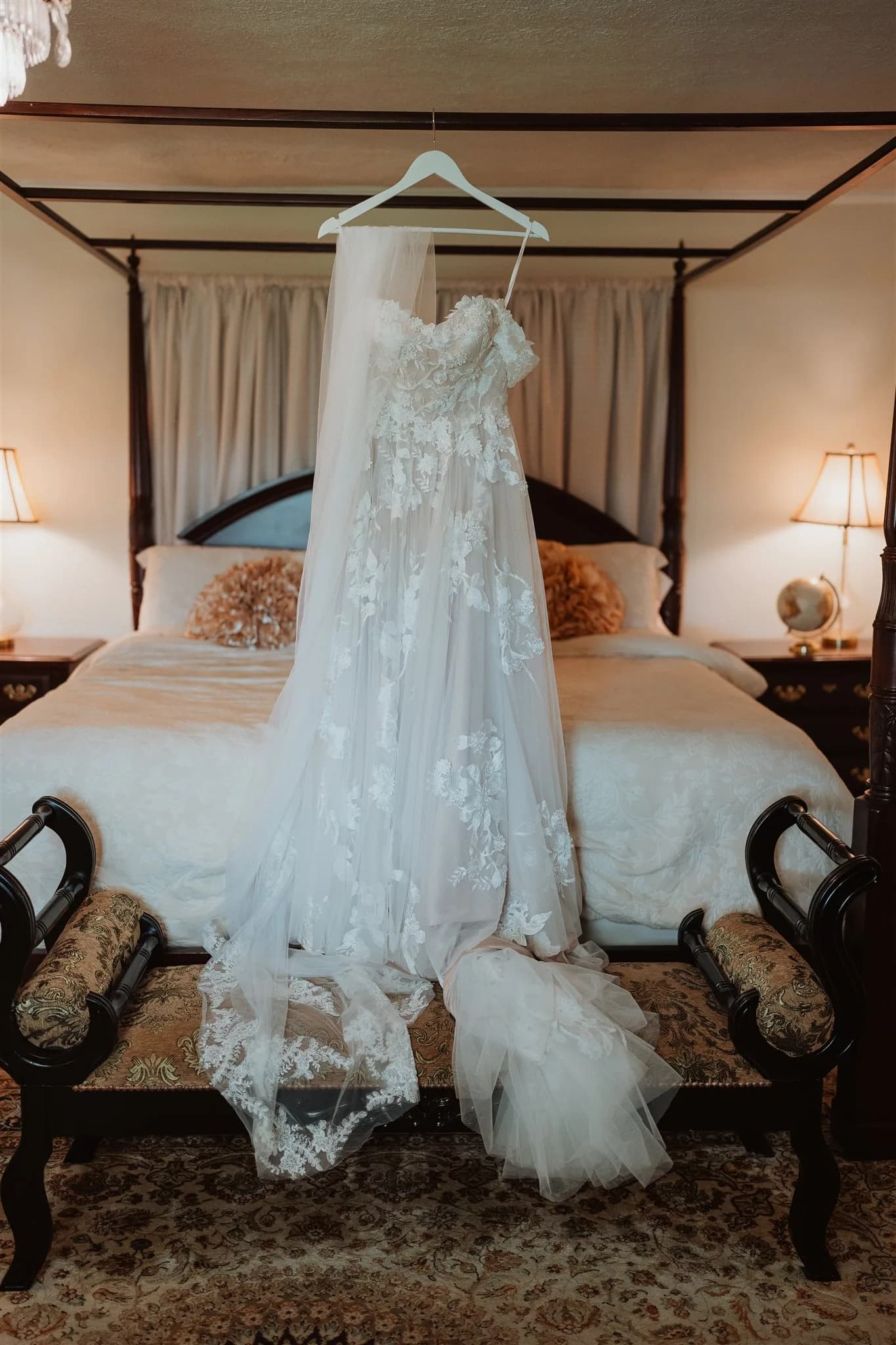 Lace wedding gown with cathedral veil hanging from four-poster bed in a warmly lit bridal suite