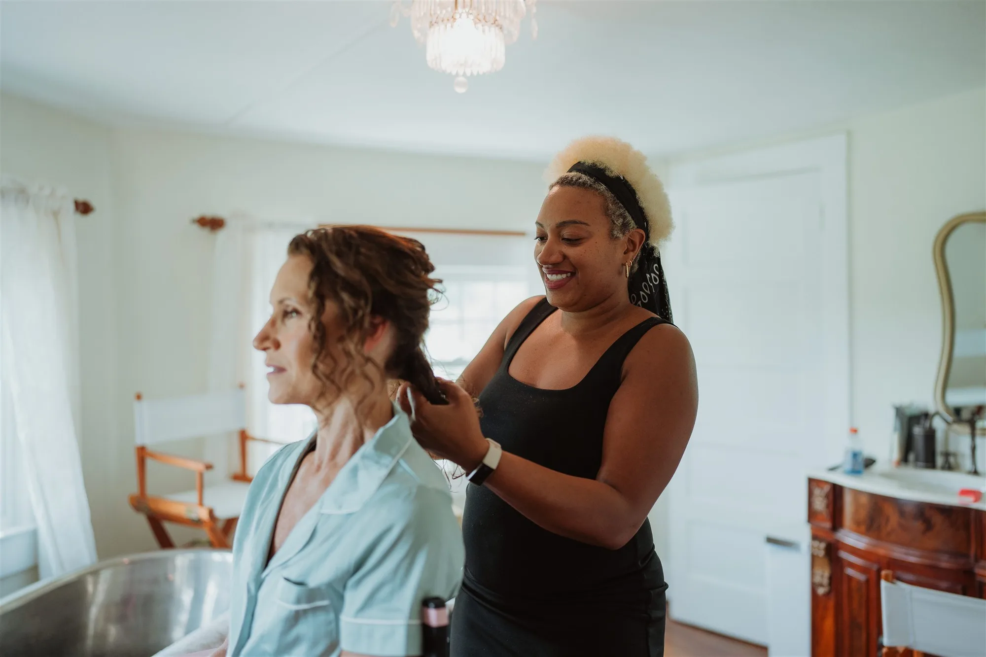 Smiling hair stylist works on bride's curls in a bright getting-ready room at Rixey Manor