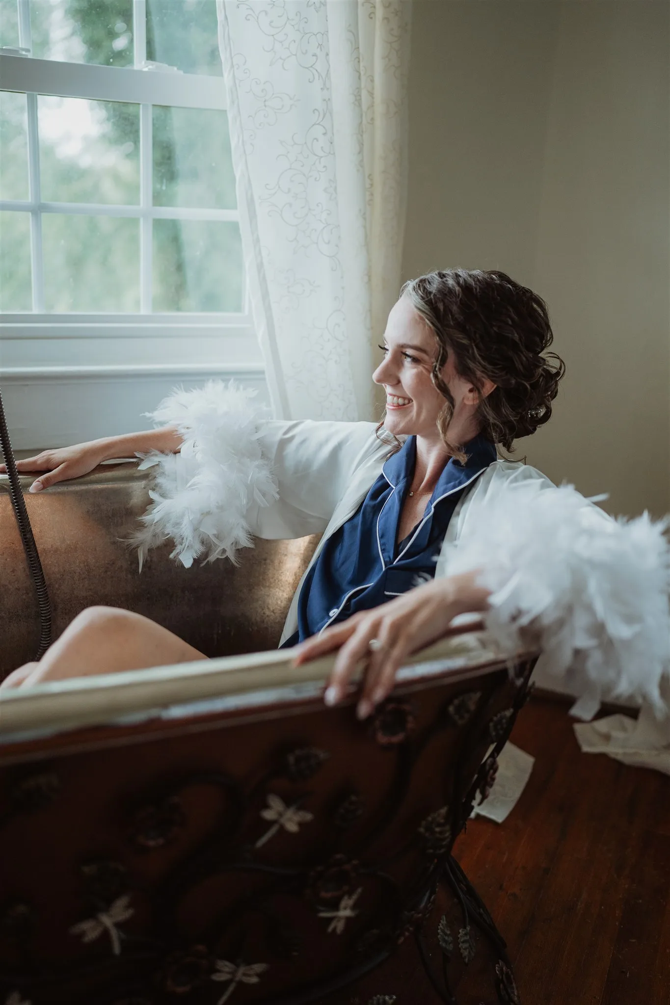 Smiling bride in feather robe relaxing in a vintage clawfoot tub by a window at Rixey Manor