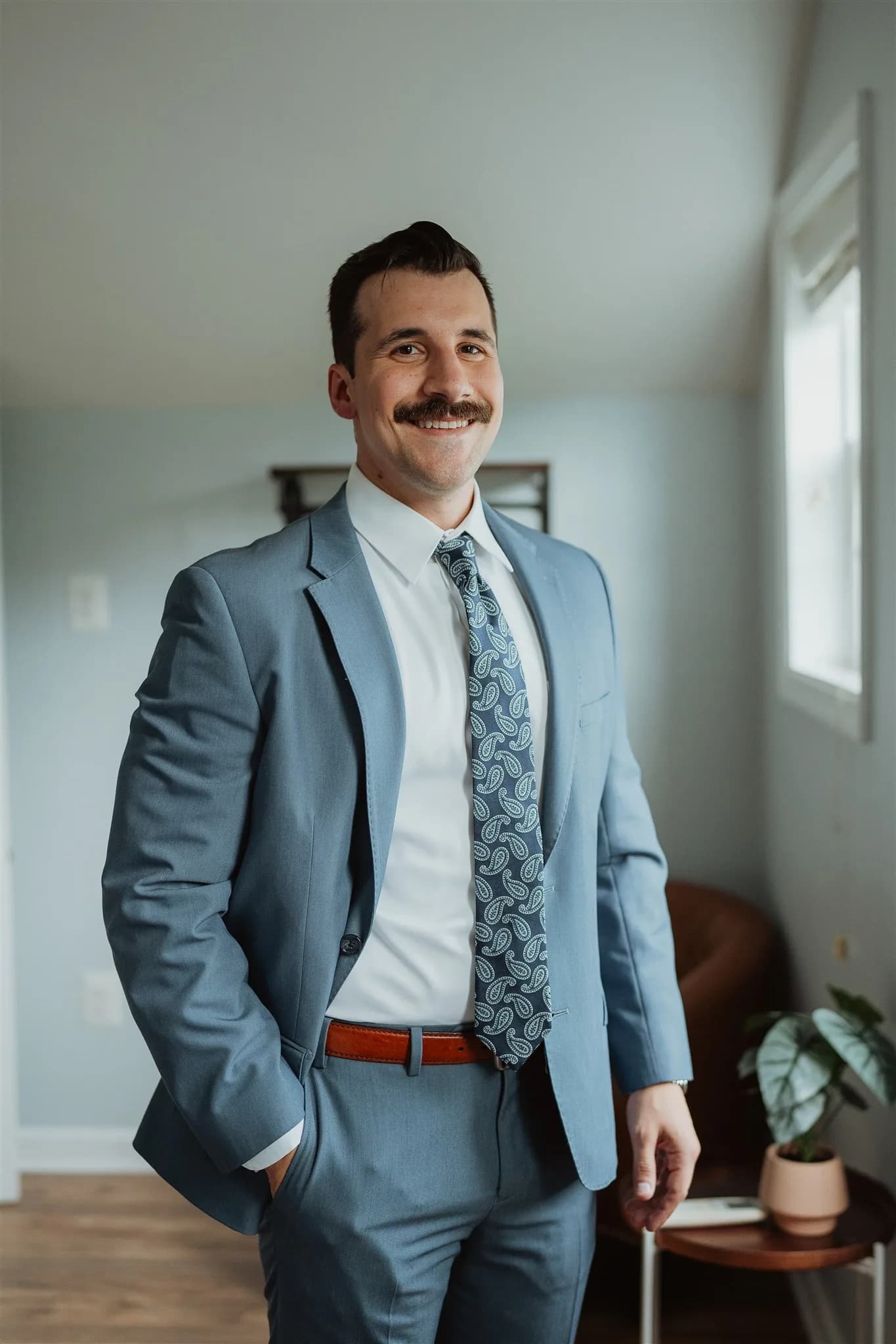 Smiling groom in light blue suit and paisley tie poses confidently indoors before the wedding ceremony
