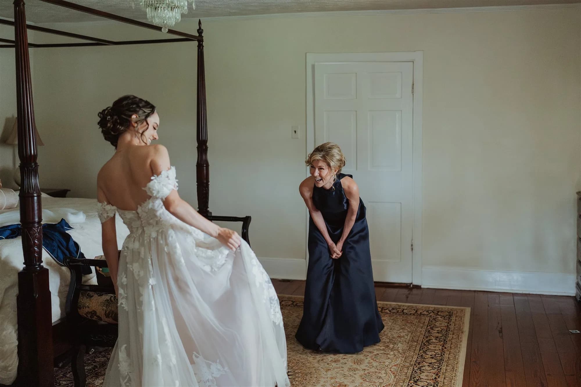 Bride in lace gown laughs with smiling bridesmaid in navy dress inside a bright bedroom at Rixey Manor