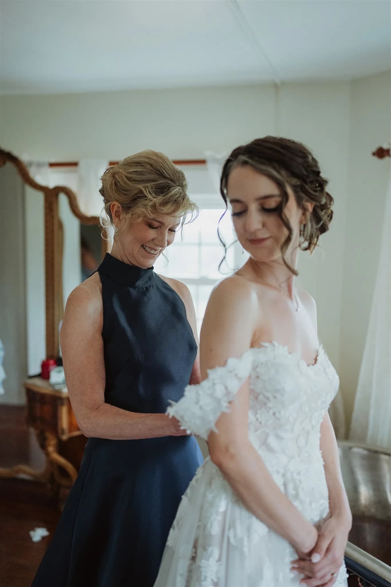 Smiling woman zips up bride's white lace gown in a warmly lit getting-ready room