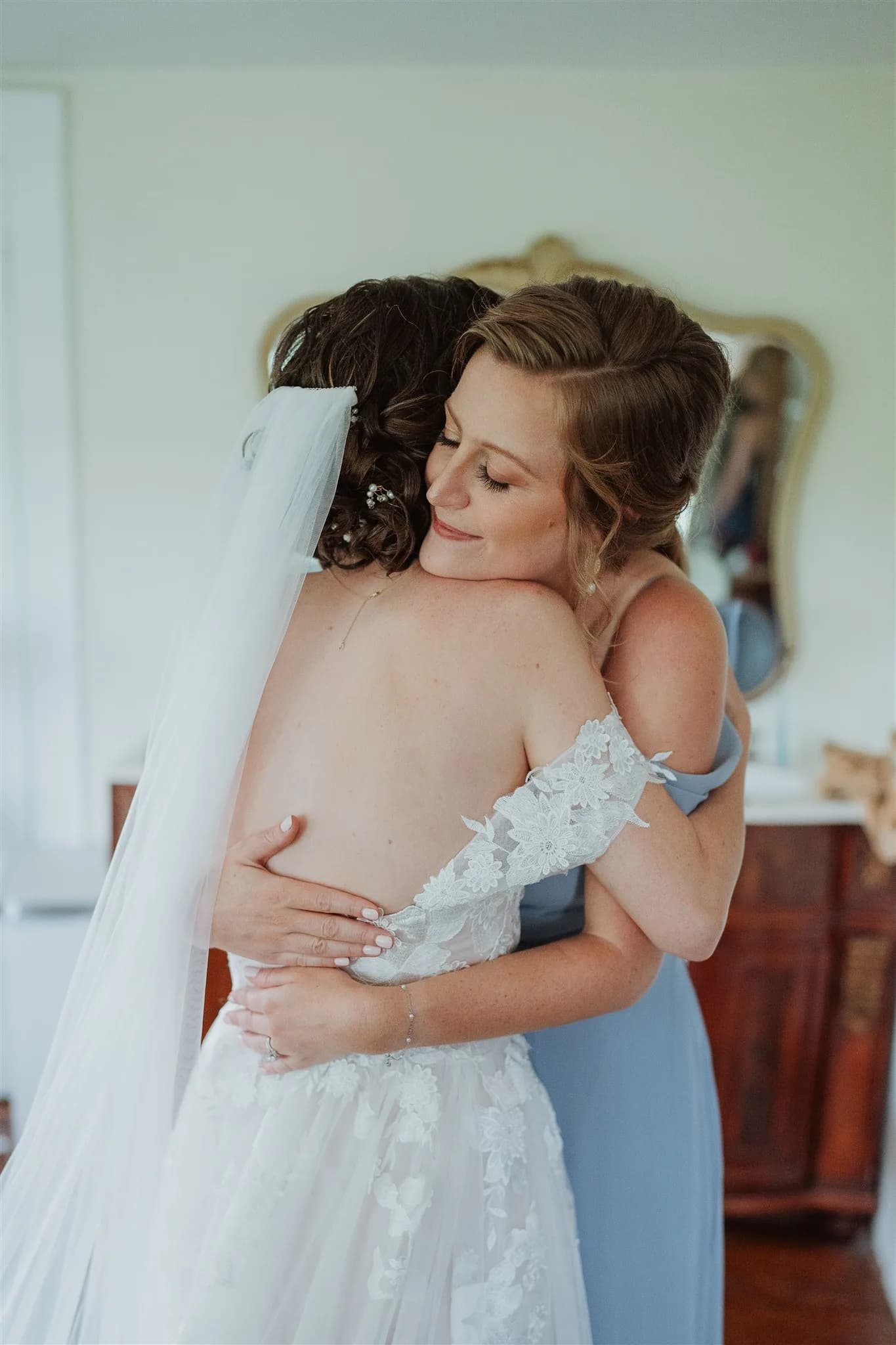 Bride in lace gown shares a tender embrace with a bridesmaid in blue during wedding morning preparations