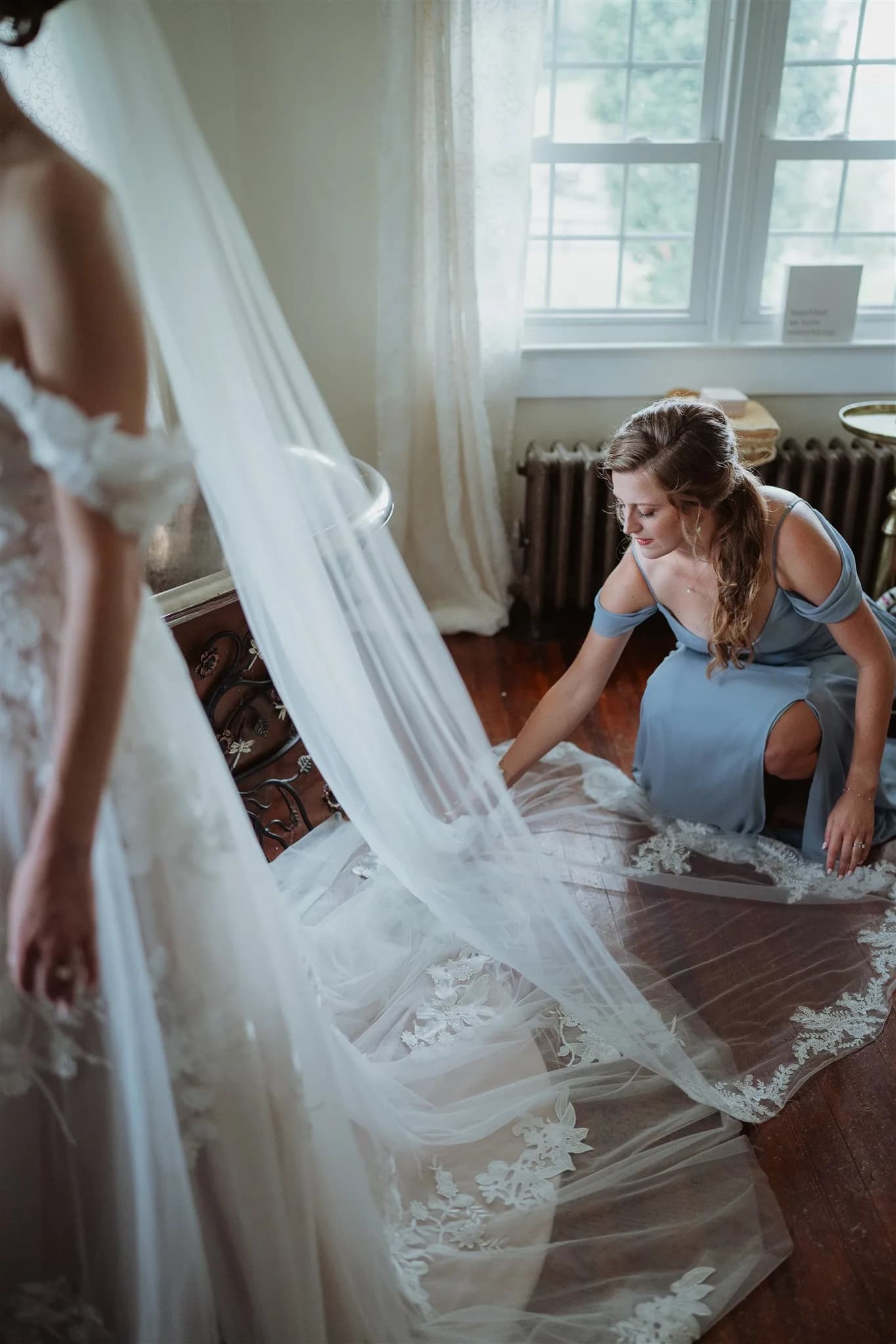 Bridesmaid in blue dress arranges lace veil on floor while bride stands nearby in sunlit room
