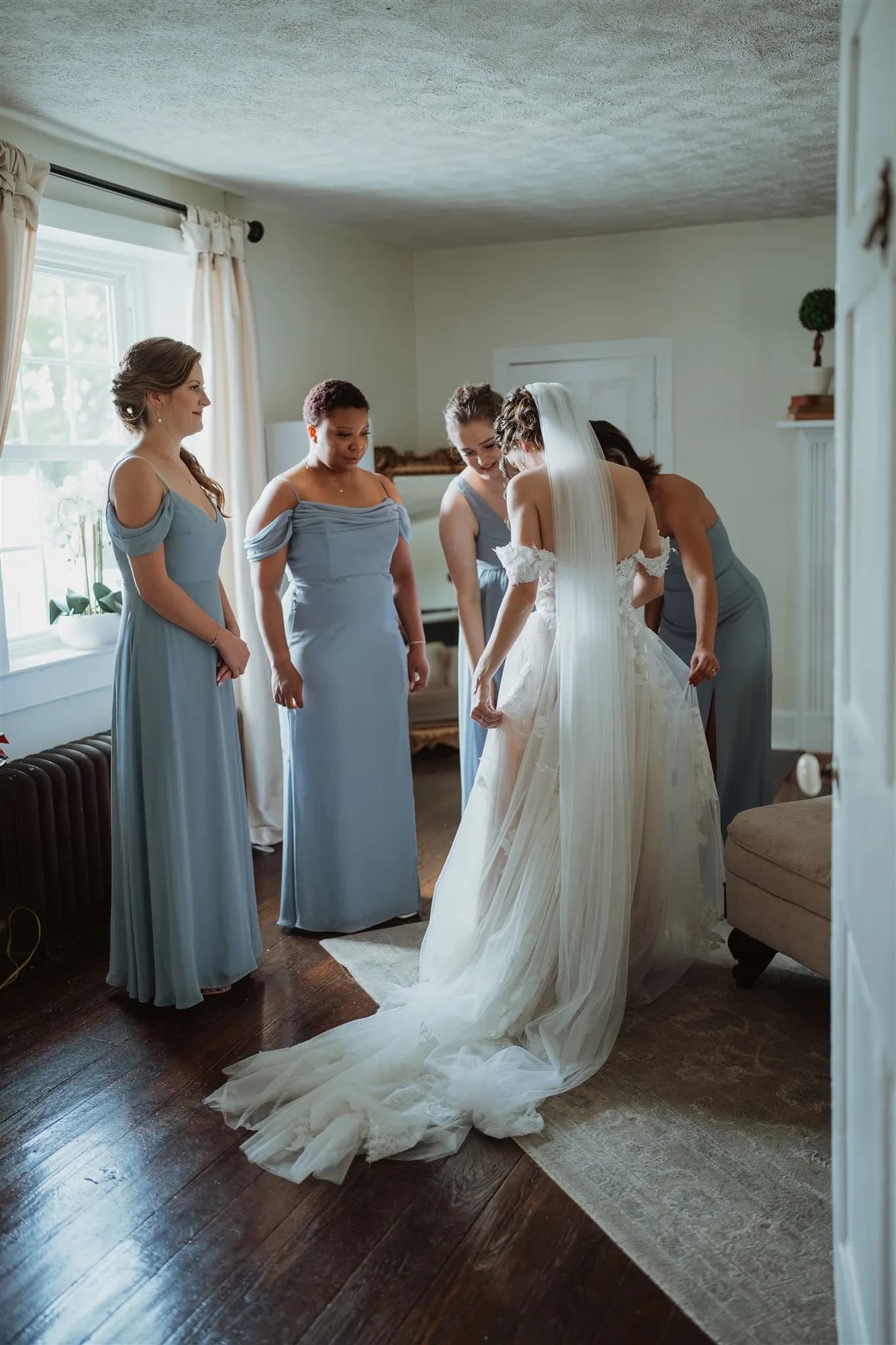 Bride in white gown and veil surrounded by bridesmaids in dusty blue dresses during getting-ready moment